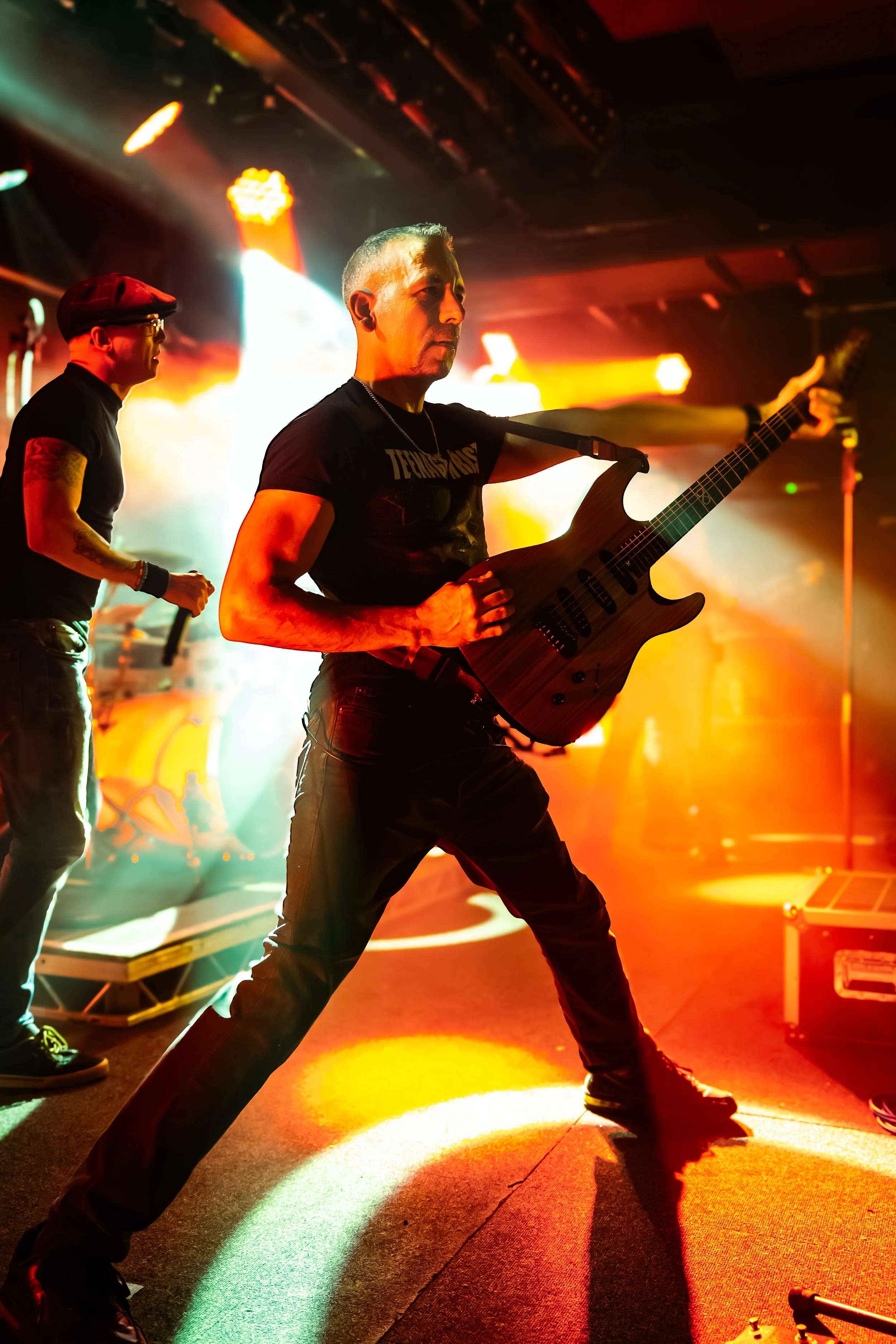 A live music performance scene with two musicians on stage: one with a guitar, striking a dynamic pose, and another with a microphone. The stage is illuminated by vibrant, multicolored lights creating an energetic atmosphere.
