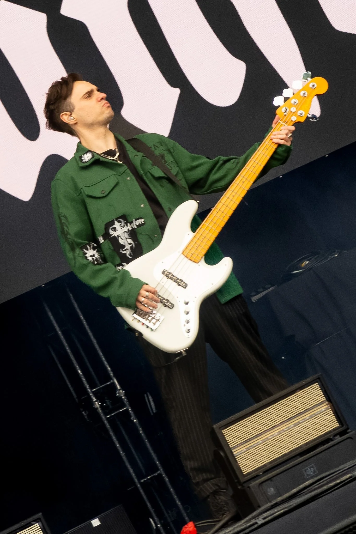 A musician wearing a green jacket playing a white electric bass guitar on stage, with a large black and white graphic background.
