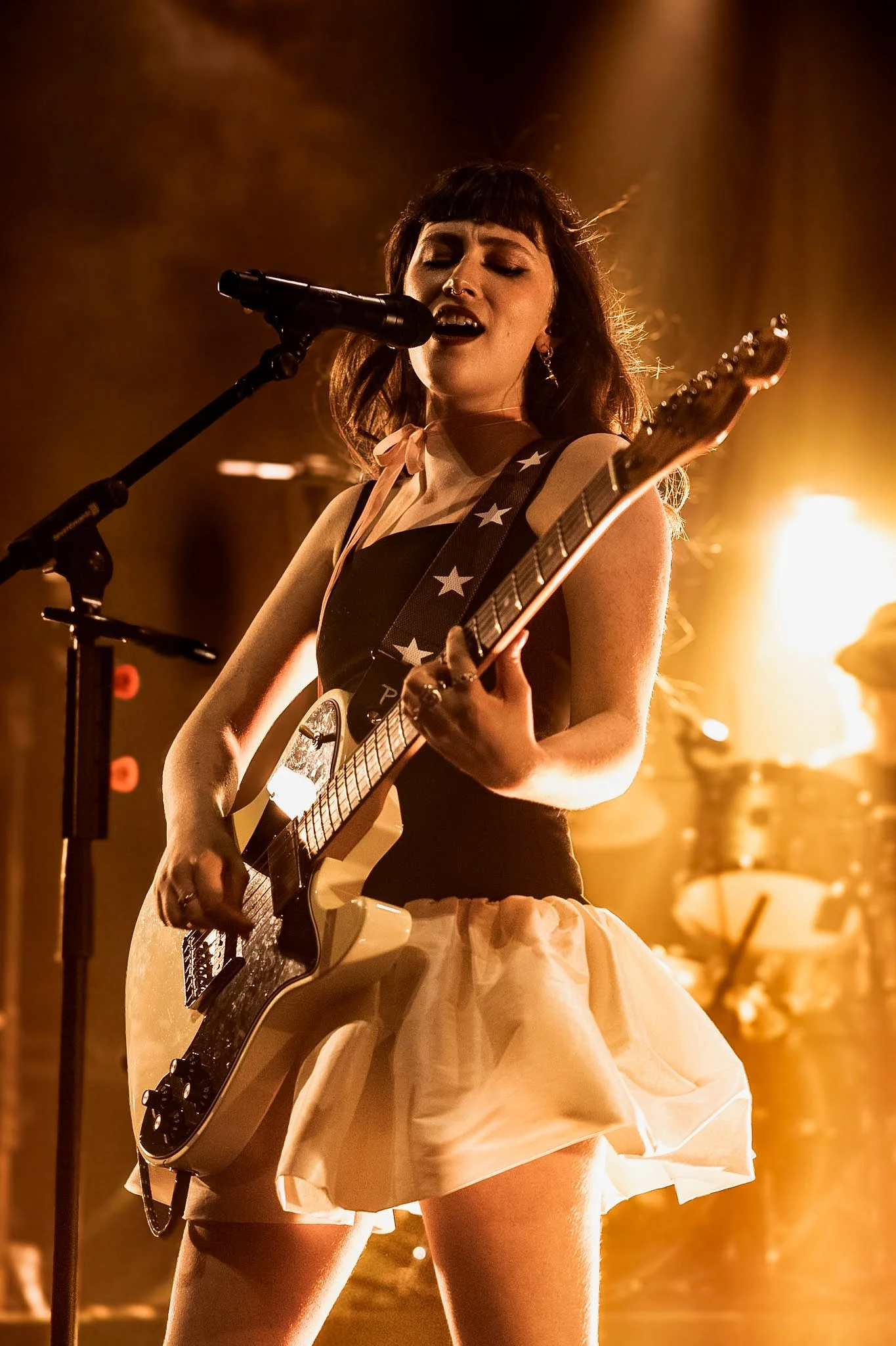 A female musician singing into a microphone while playing an electric guitar on stage, illuminated by warm stage lighting.