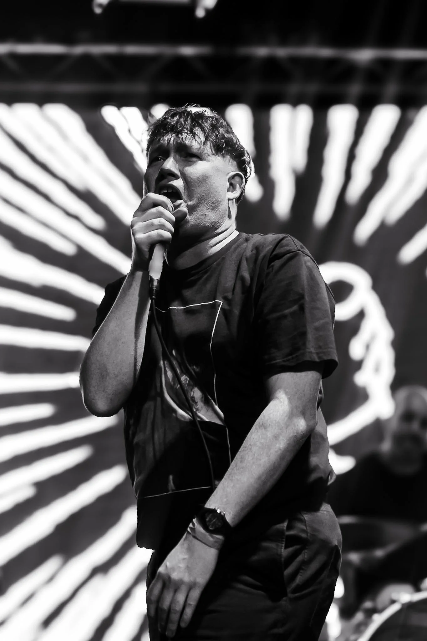 Black and white photo of a man singing into a microphone with expressive face and messy hair. He is wearing a t-shirt and a watch, with a background of illuminated stripes.