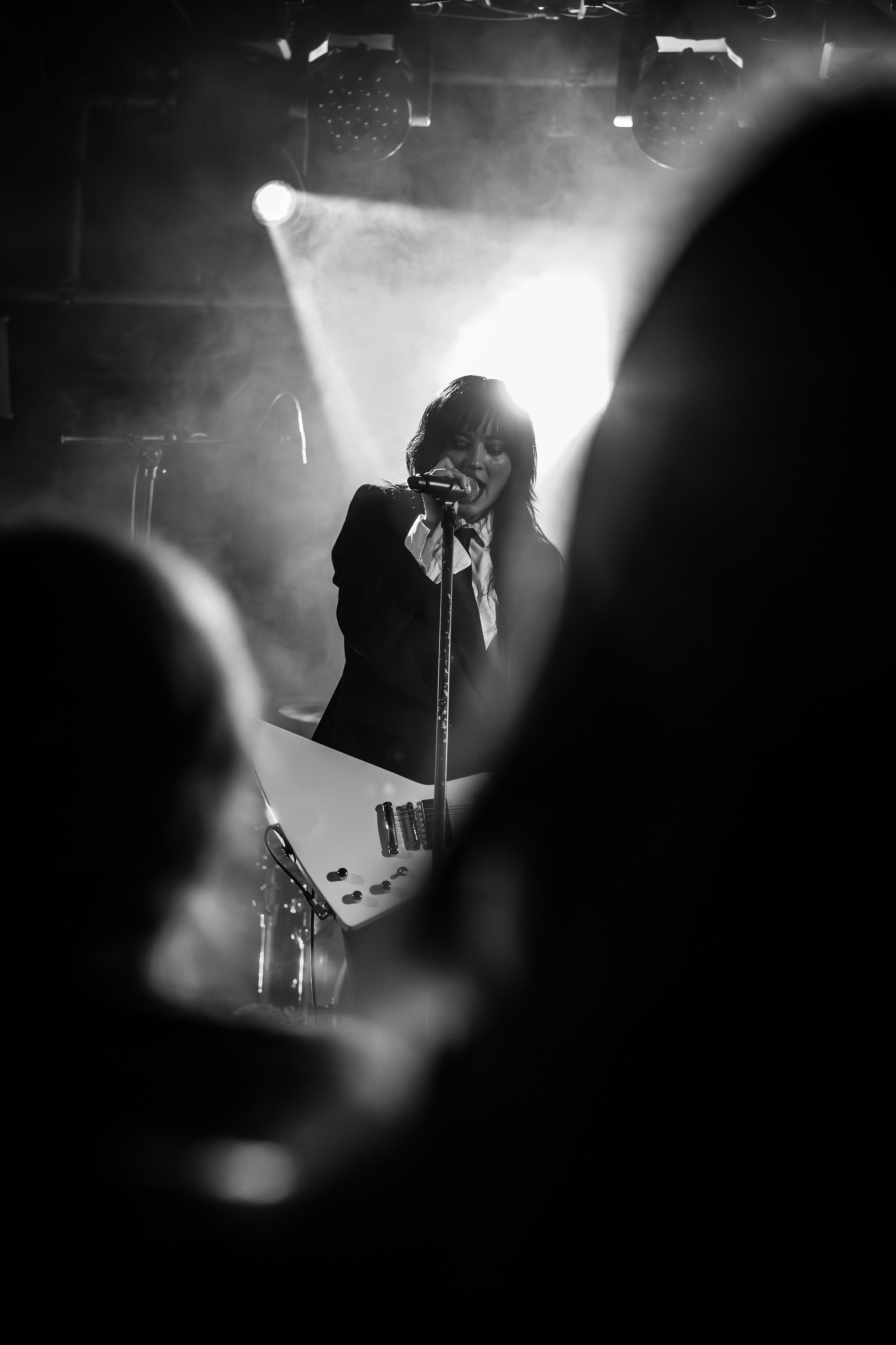 A female singer performs with a guitar on stage, illuminated by a bright light behind her, in a black and white photo. The audience is visible as dark silhouettes in the foreground.