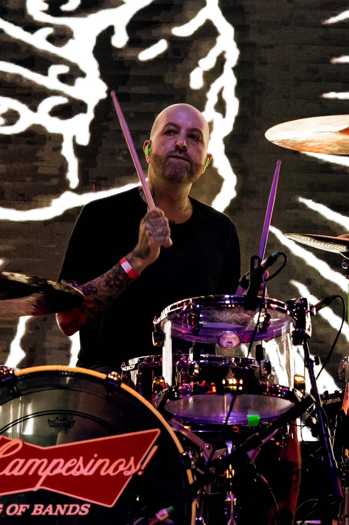 A man playing drums on stage with a large black and white patterned background behind him, holding drumsticks, wearing a black shirt, and having a beard and shaved head.