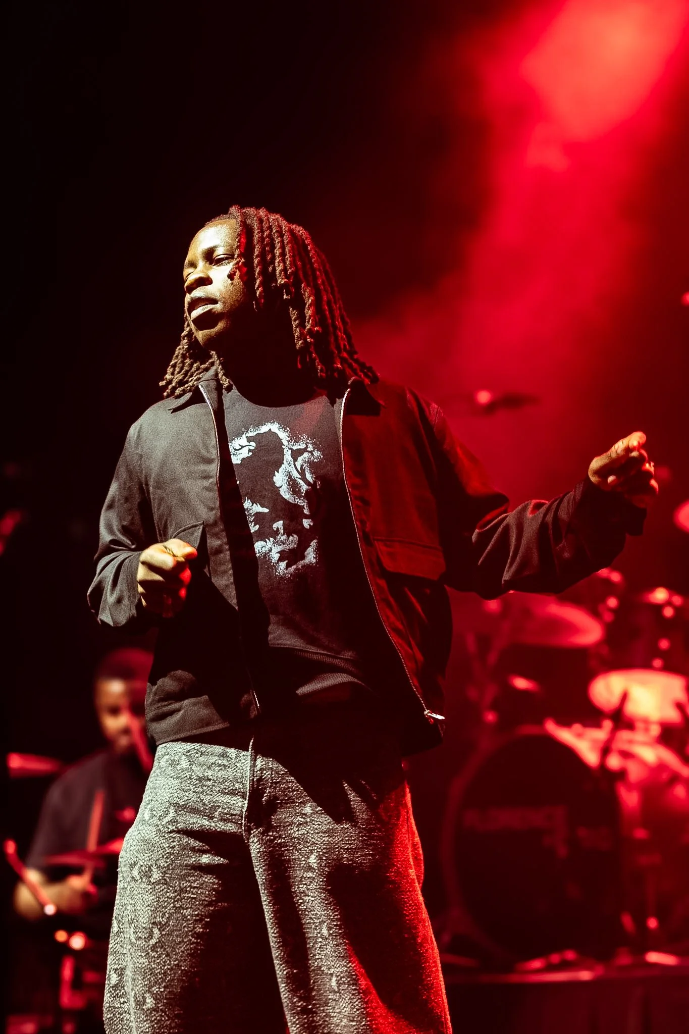 Performer with red dreadlocks on stage, wearing a black jacket and gray pants, holding a microphone, with a band playing in the background under red stage lighting.