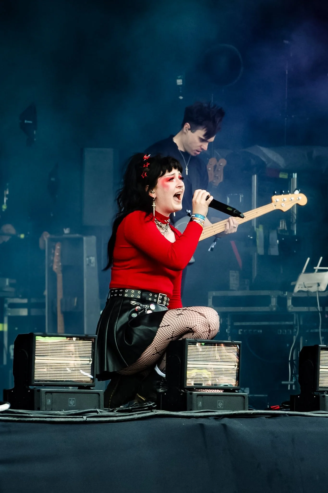 A performer singing into a microphone on stage with a guitarist in the background, colorful stage lighting highlighting their expressions.