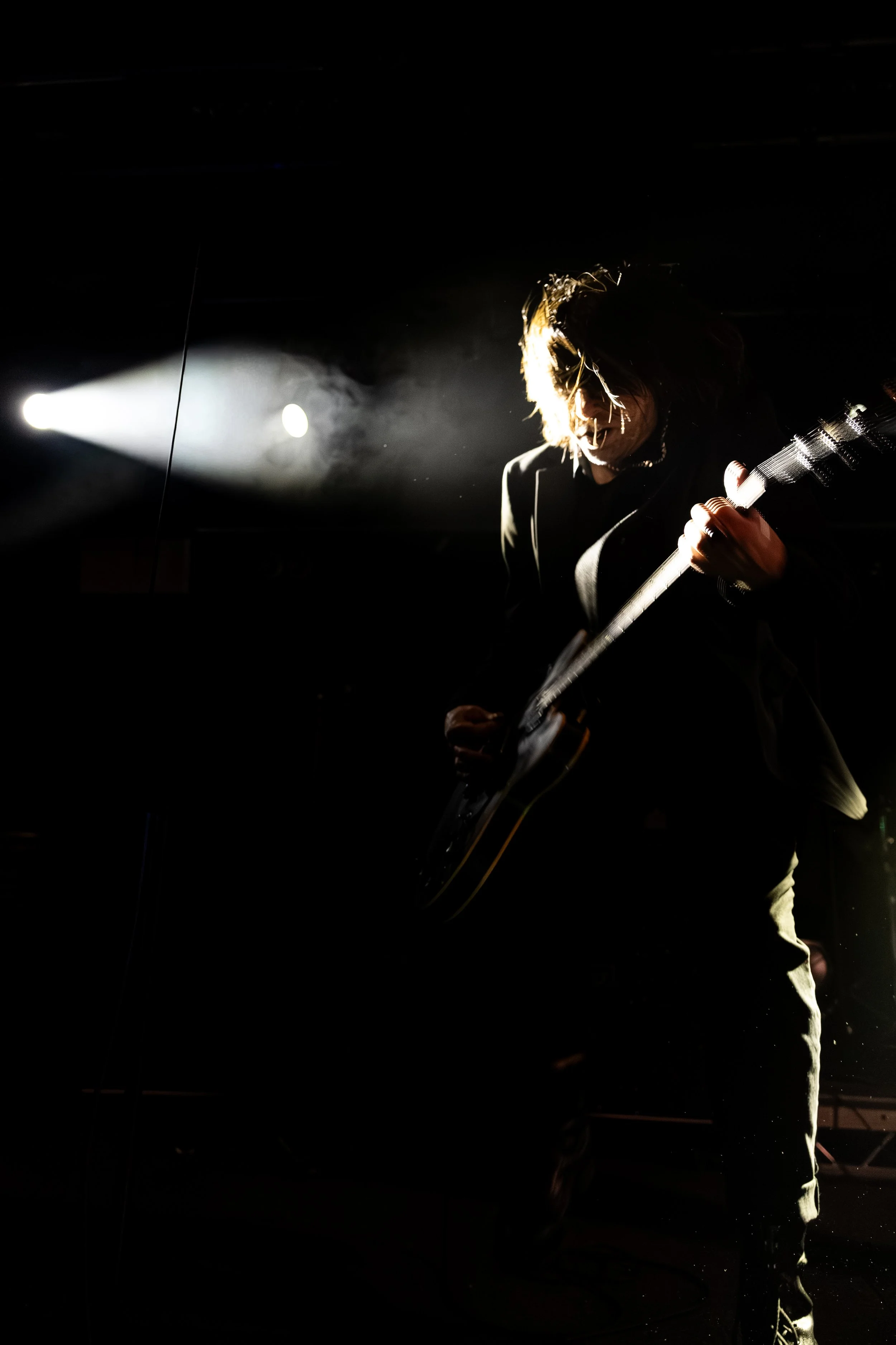 A person playing an electric guitar on stage under a bright spotlight in a dark setting.