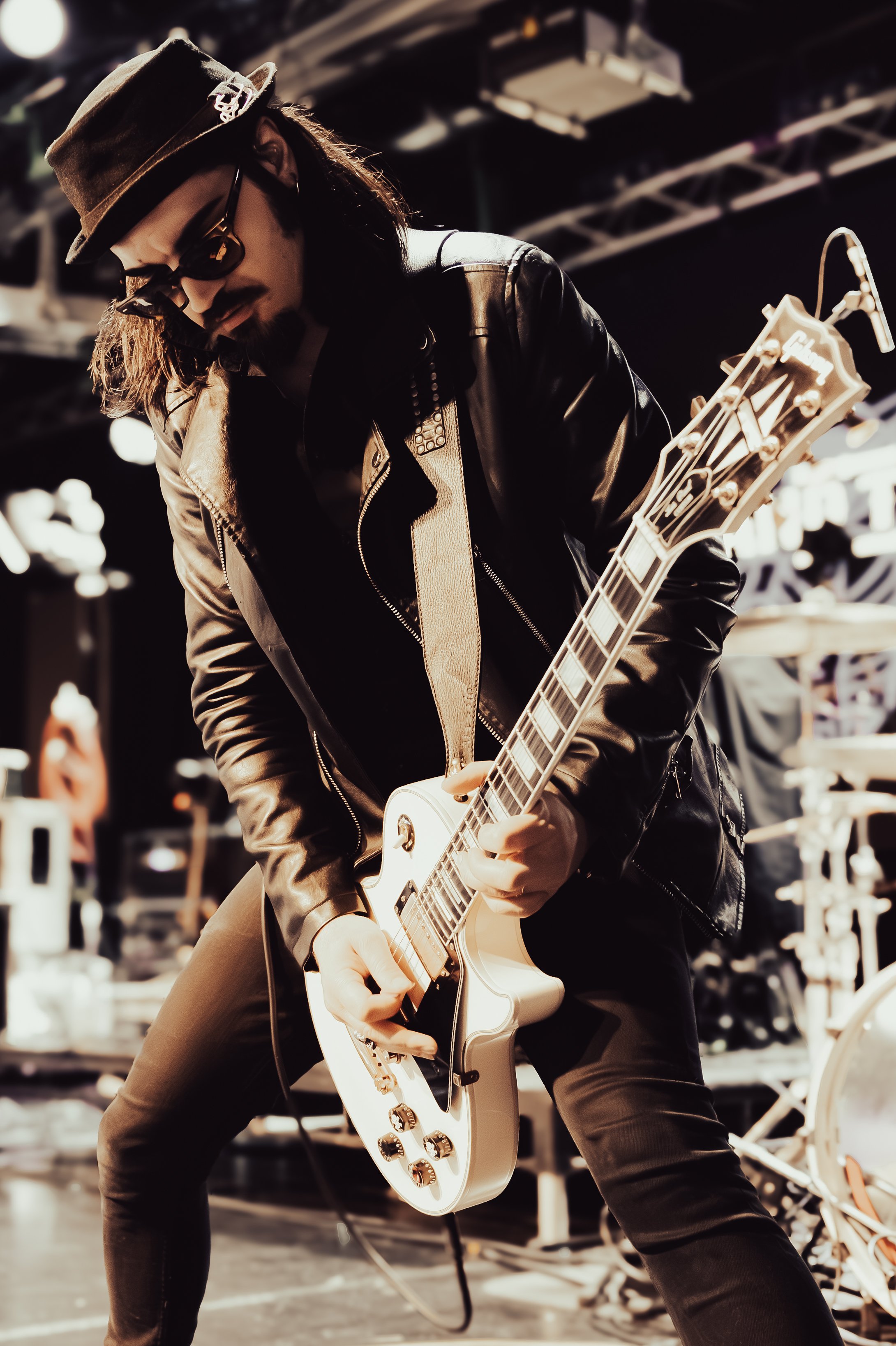 A musician wearing a leather jacket, dark sunglasses, a hat, and earrings is playing an electric guitar on stage with a blurred background of stage equipment.