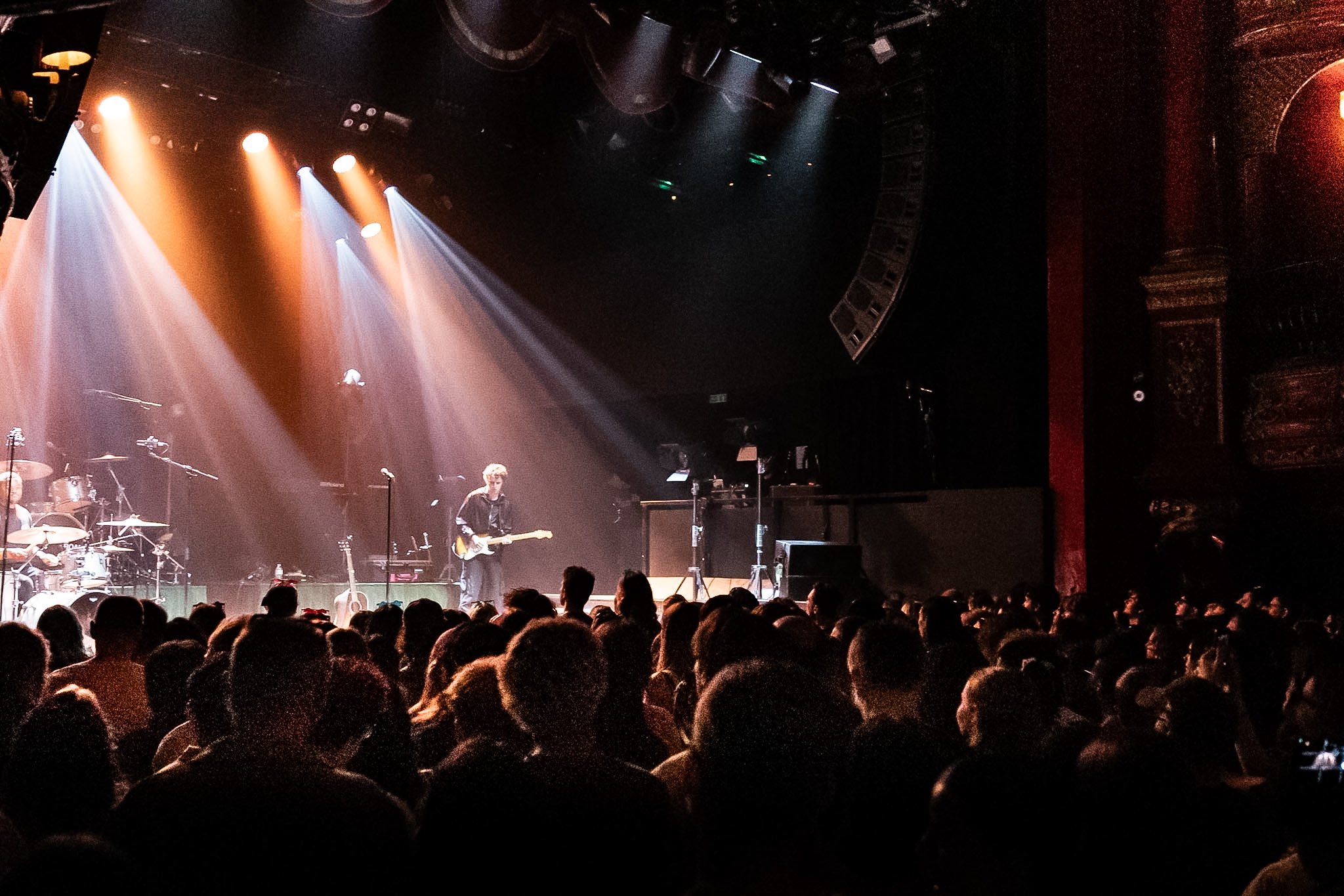 A concert stage with musicians performing under orange, white, and purple stage lights, and a large audience in the foreground.