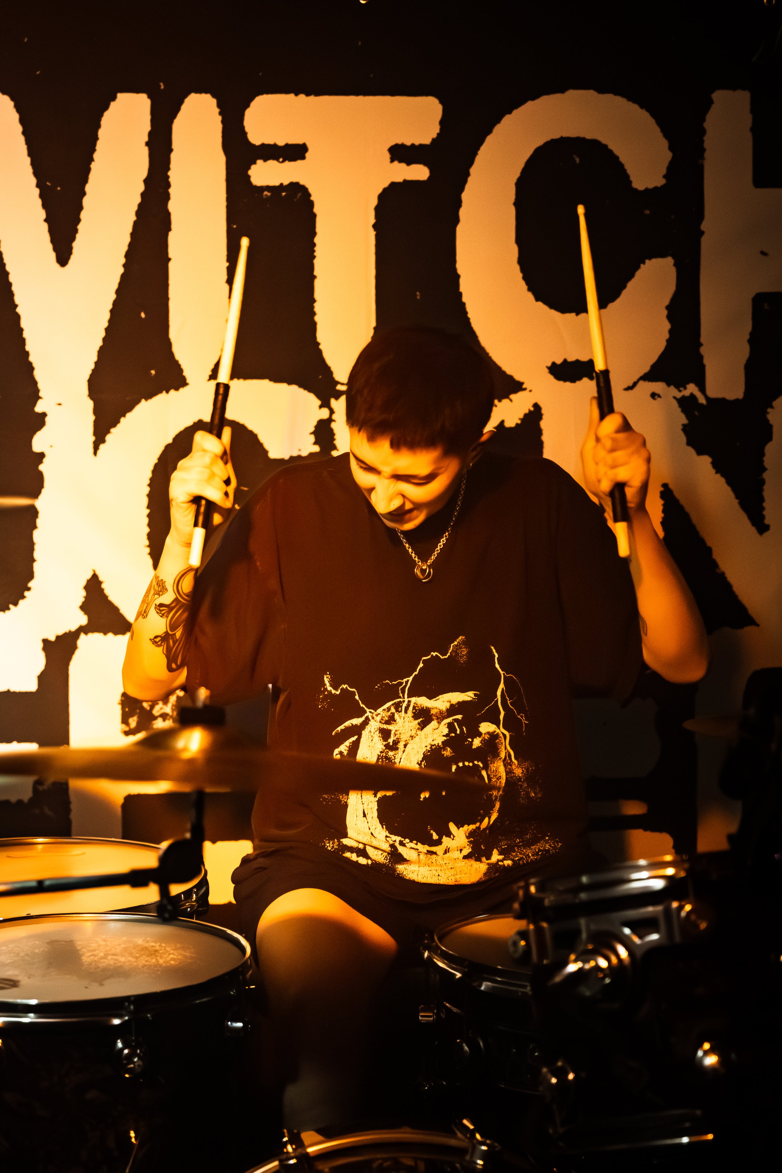 A person playing drums in a dimly lit room with the word 'WET' in large letters on the wall behind them. The person is wearing a black t-shirt with a skull graphic and has short hair and arm tattoos.