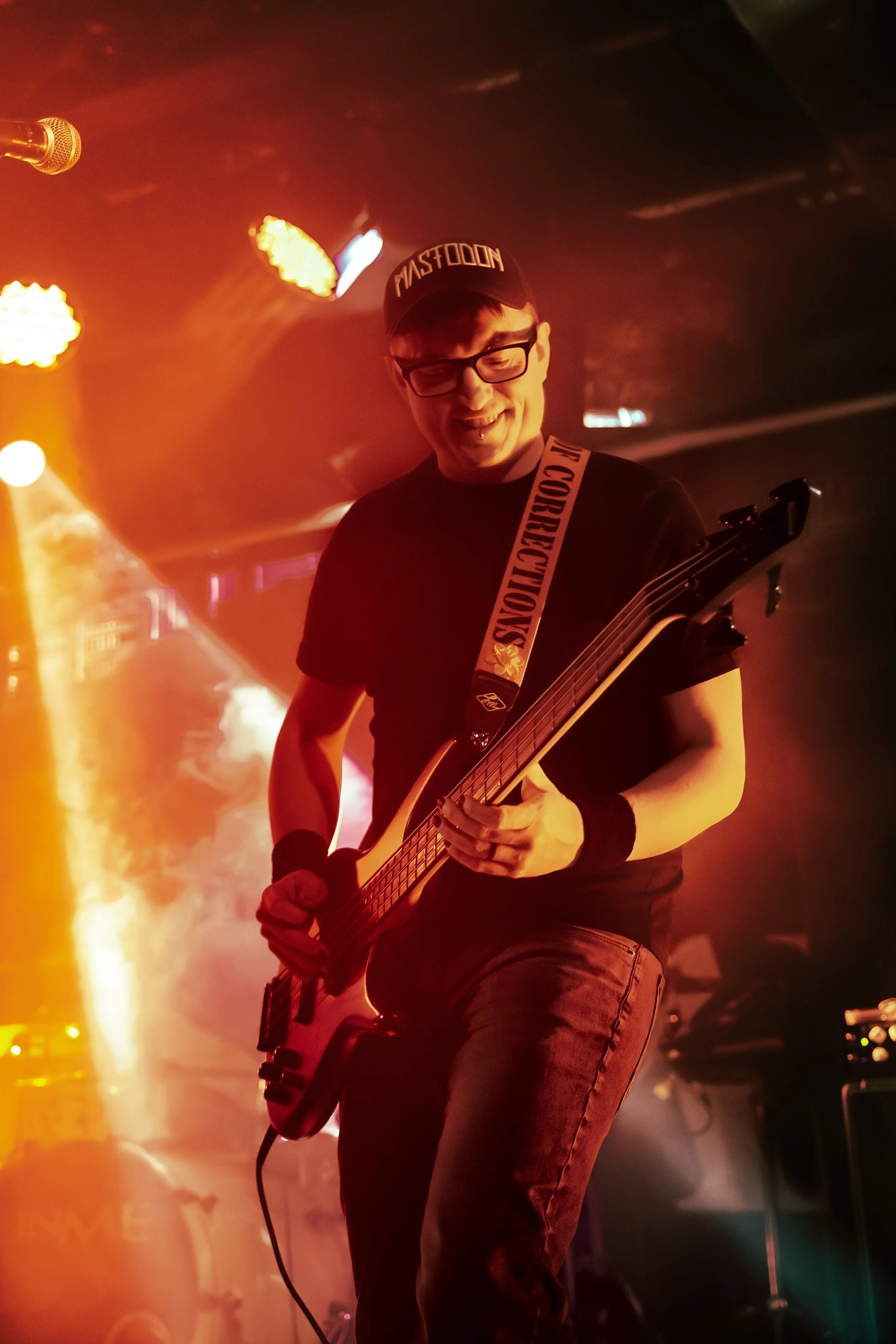 A man wearing glasses and a black cap playing an electric guitar on stage amid red and yellow stage lighting.