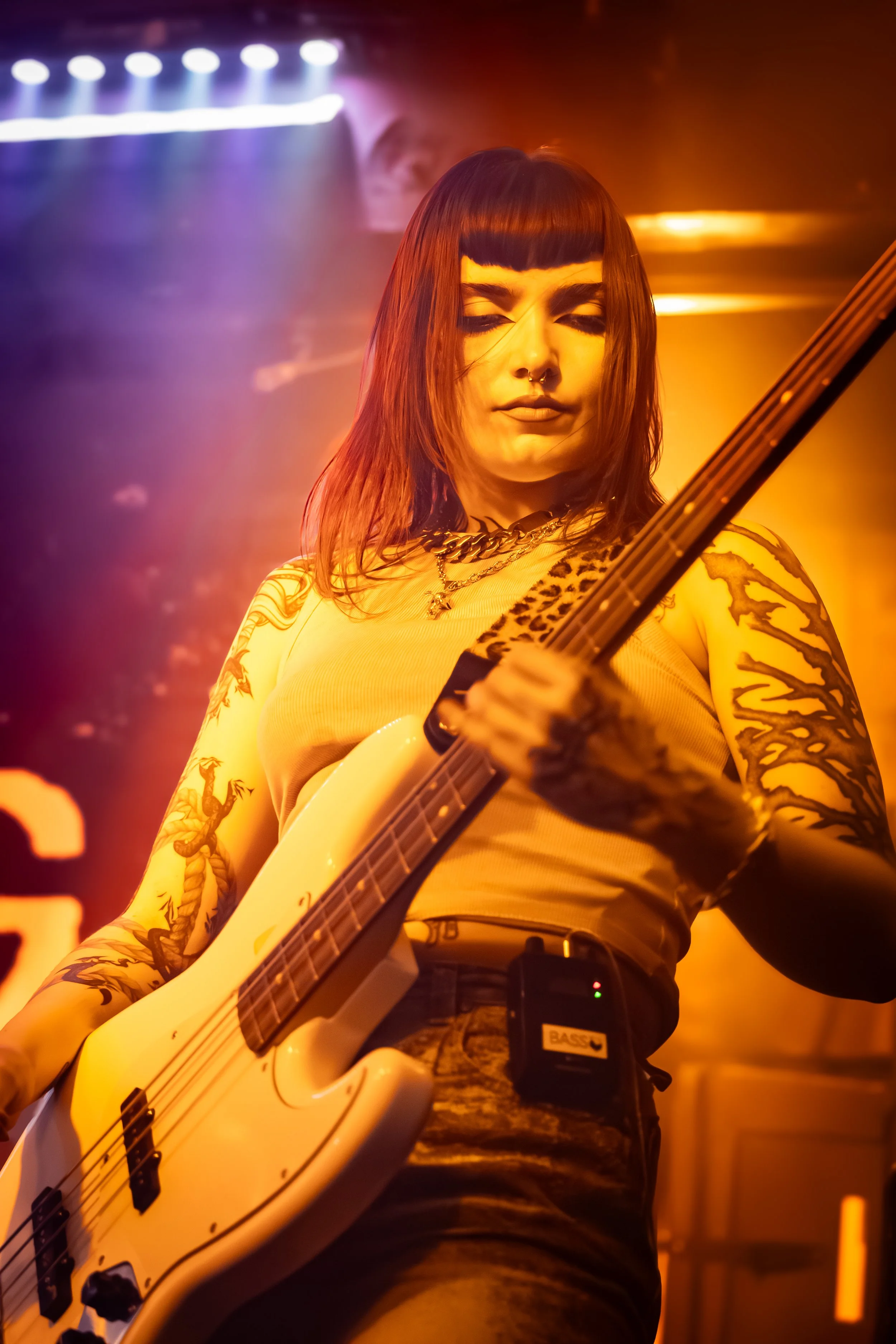 A woman playing an electric guitar on stage, with multicolored stage lights in background, wearing a yellow top, with dark hair and tattoos on her arms.