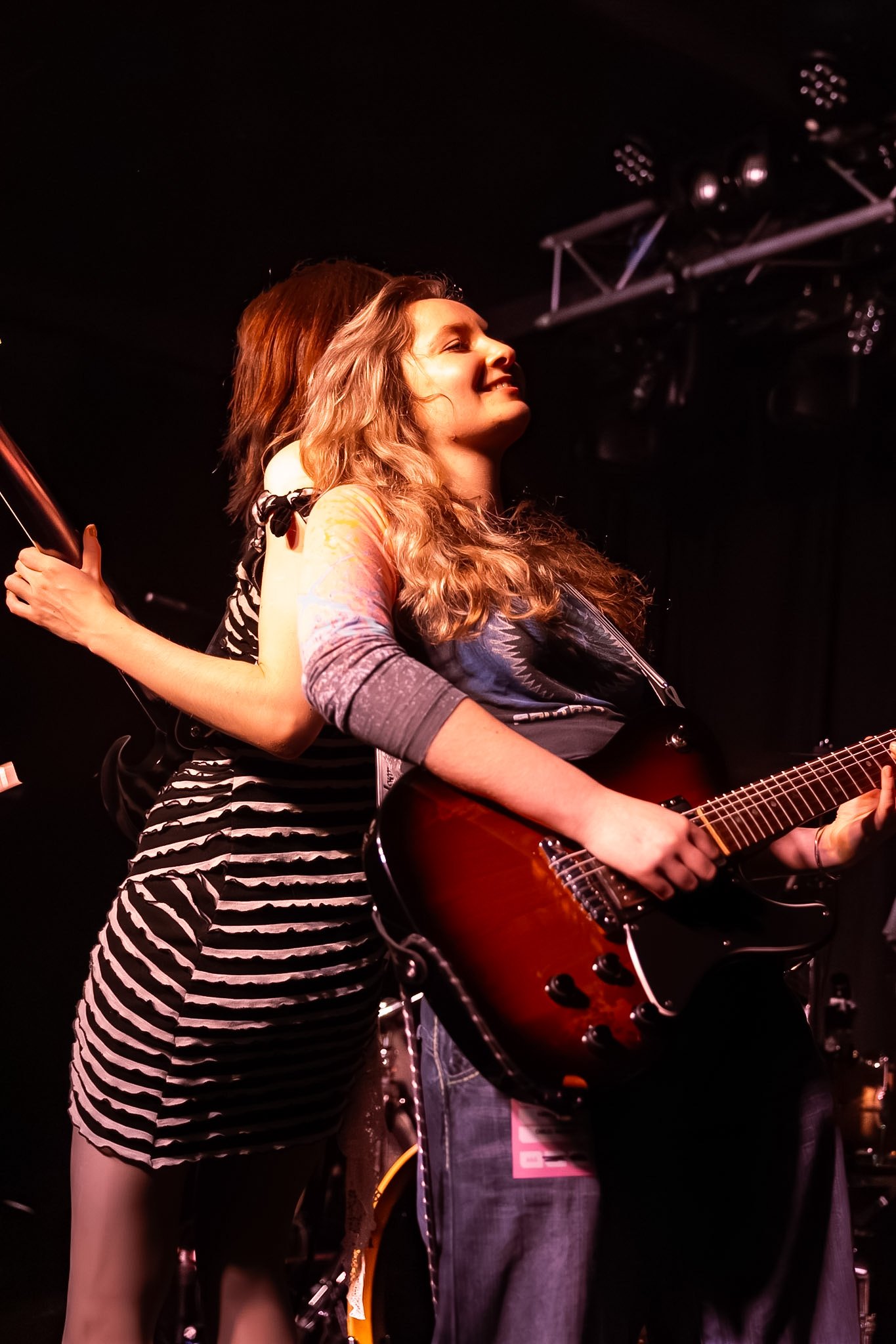 Two women performing on stage, one playing guitar and the other singing, with stage lighting and equipment visible.