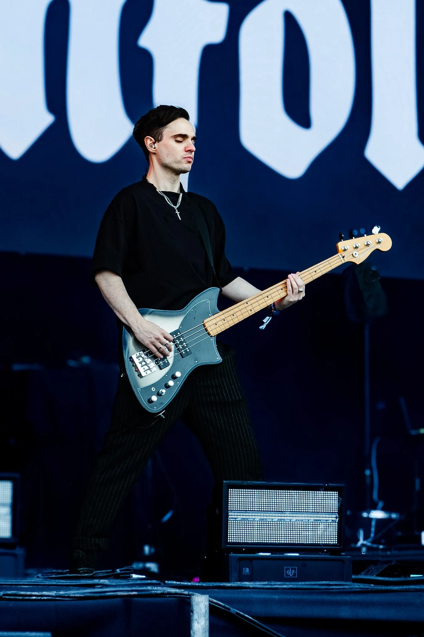A musician playing a bass guitar on stage during a performance with a large abstract backdrop in the background.