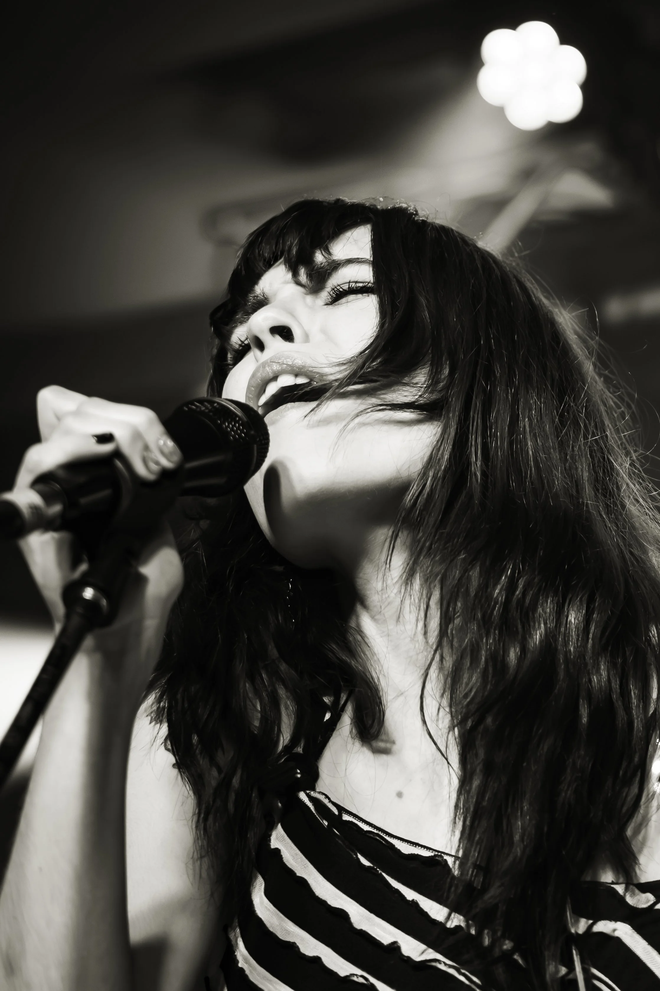 Black and white photograph of a woman singing into a microphone. She has dark, wavy hair, and is wearing a striped top. The background is blurred with a light source visible.
