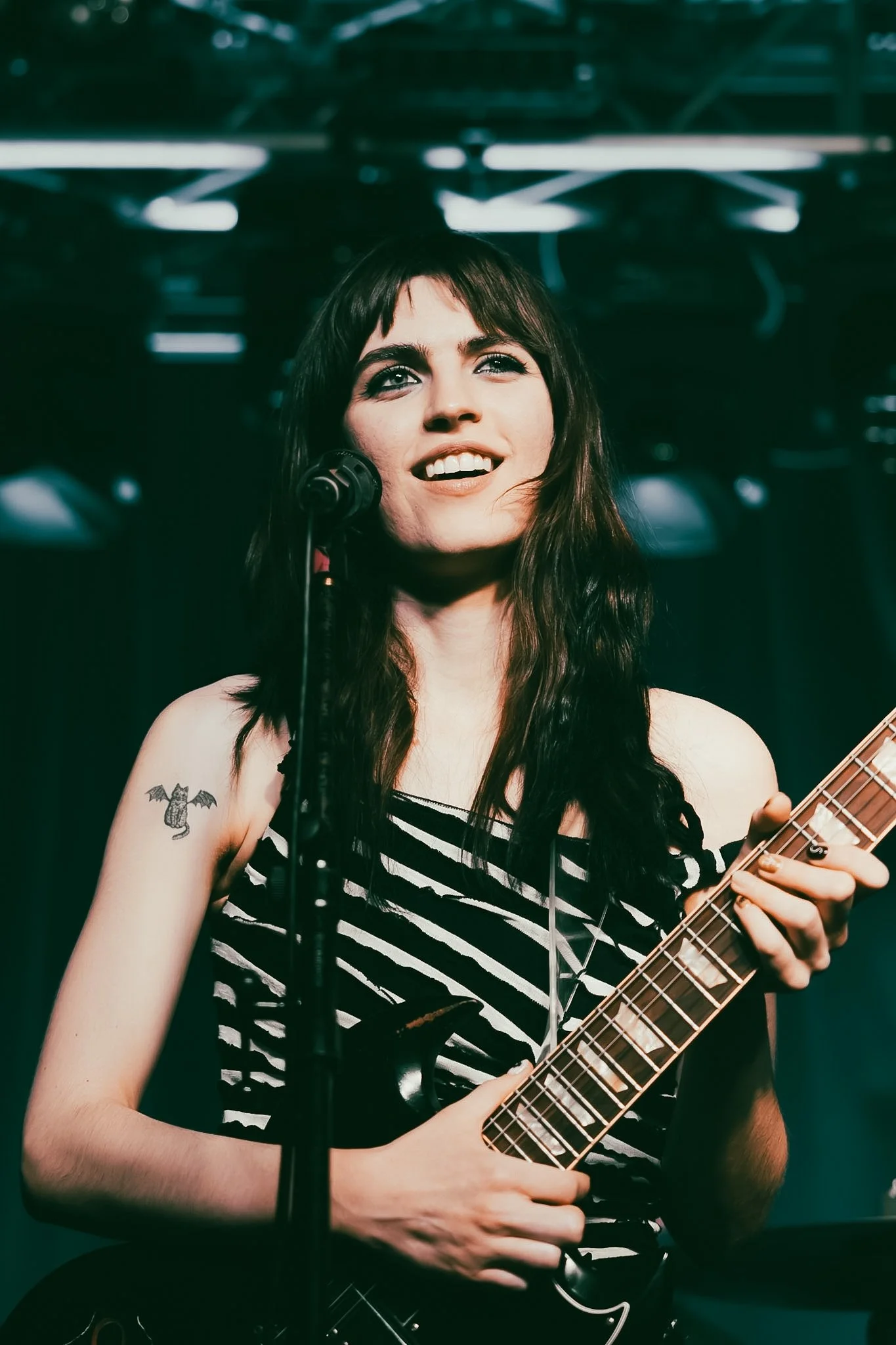 A young woman with long dark hair, tattoos on her right arm, and wearing a black and white striped top, is singing into a microphone while playing an electric guitar on stage with blue and green lighting.