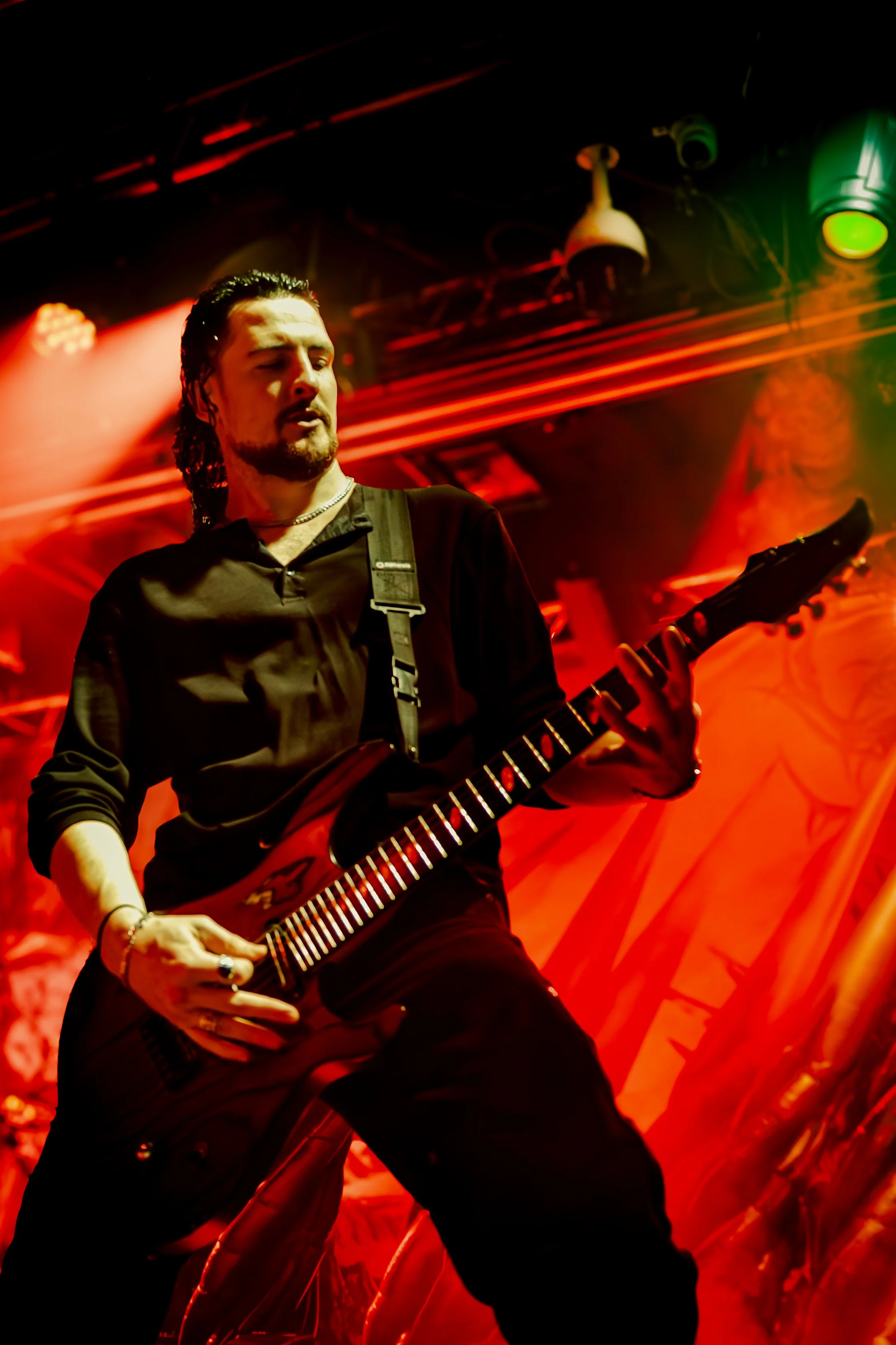 A male musician with long hair, a goatee, wearing a black shirt, playing an electric guitar on stage under red and green lighting.