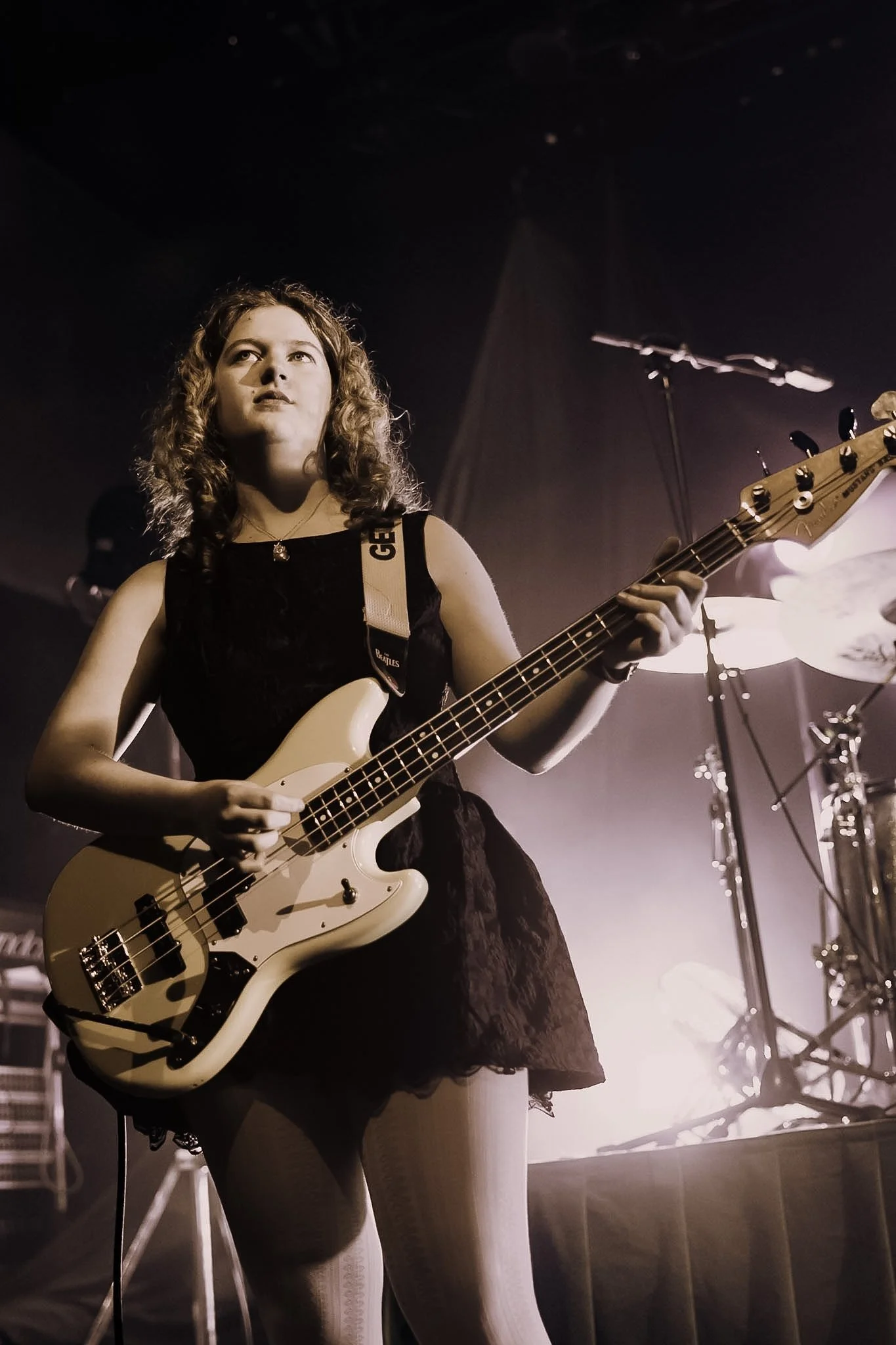 A young woman with curly hair playing an electric bass guitar on stage during a concert, illuminated by stage lights.