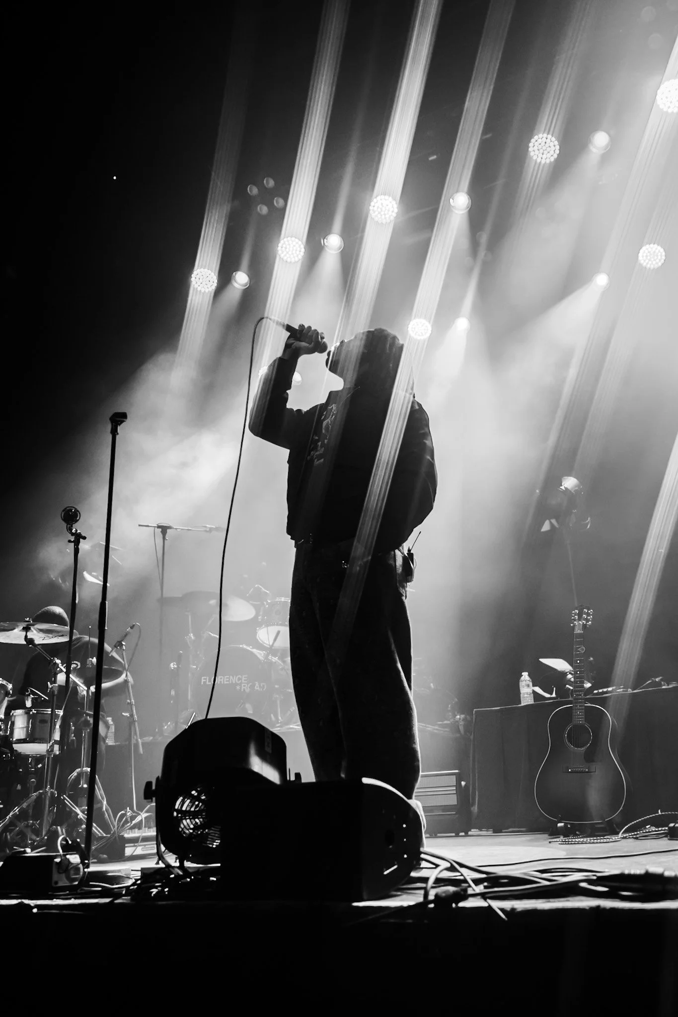 Black and white photo of a singer performing on stage with bright lights and musical instruments in the background.