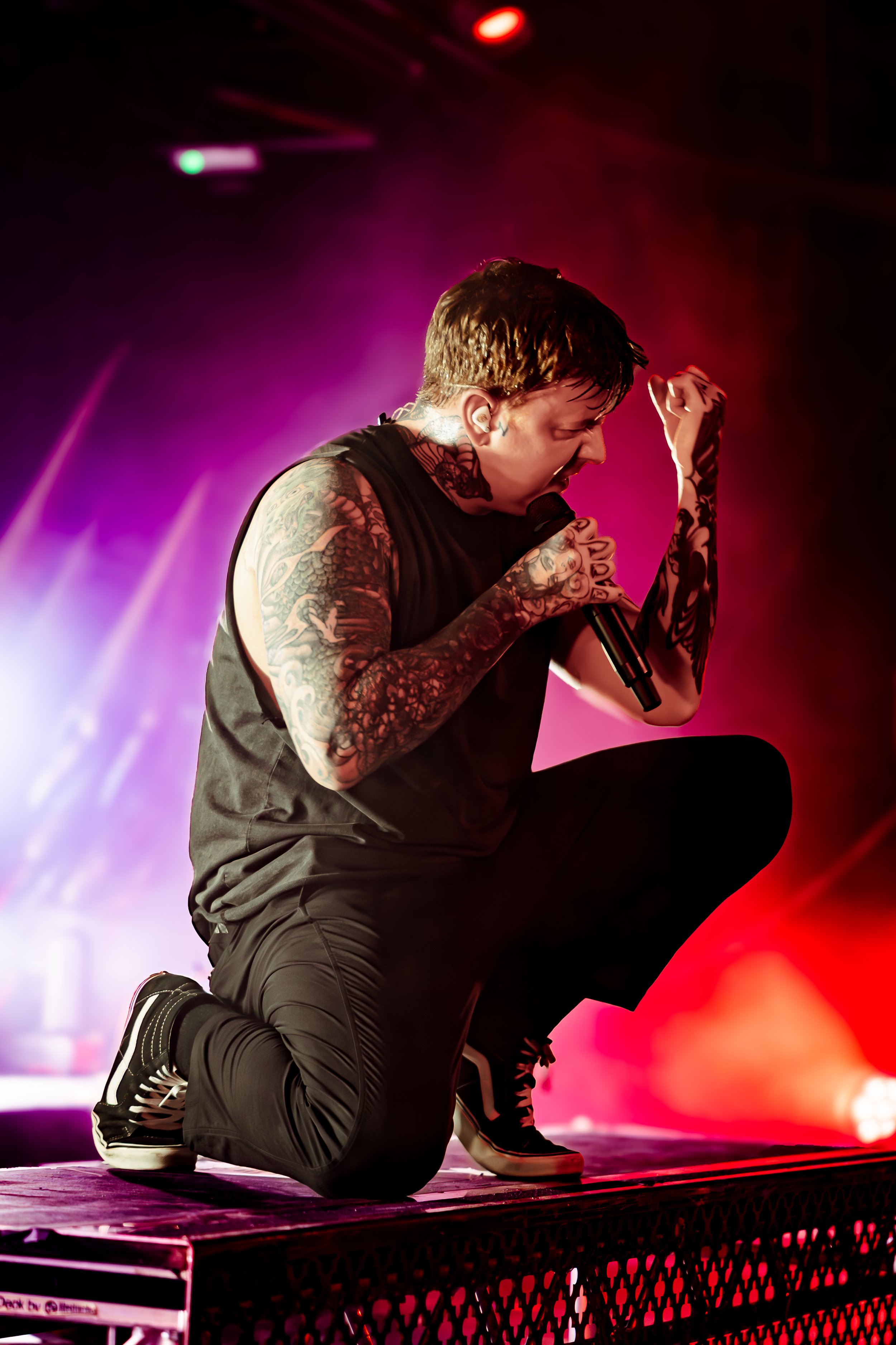 Musician with tattoos and short hair singing or shouting into a microphone on stage, kneeling on one knee with colorful stage lighting effect.