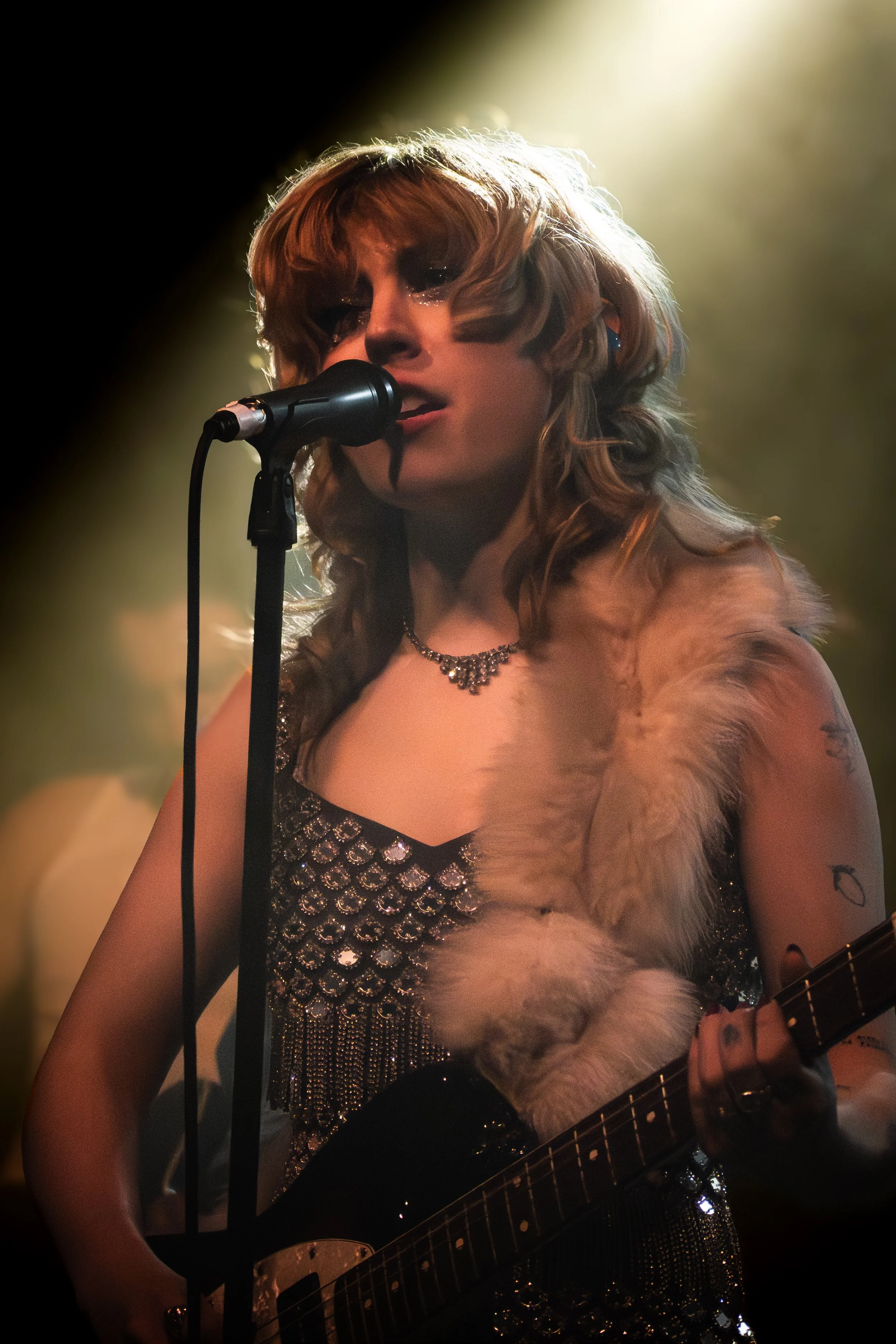 A woman singing into a microphone while playing an electric guitar, dressed in a sequined outfit and a fur stole, with lighting highlighting her face and hair during a live performance.