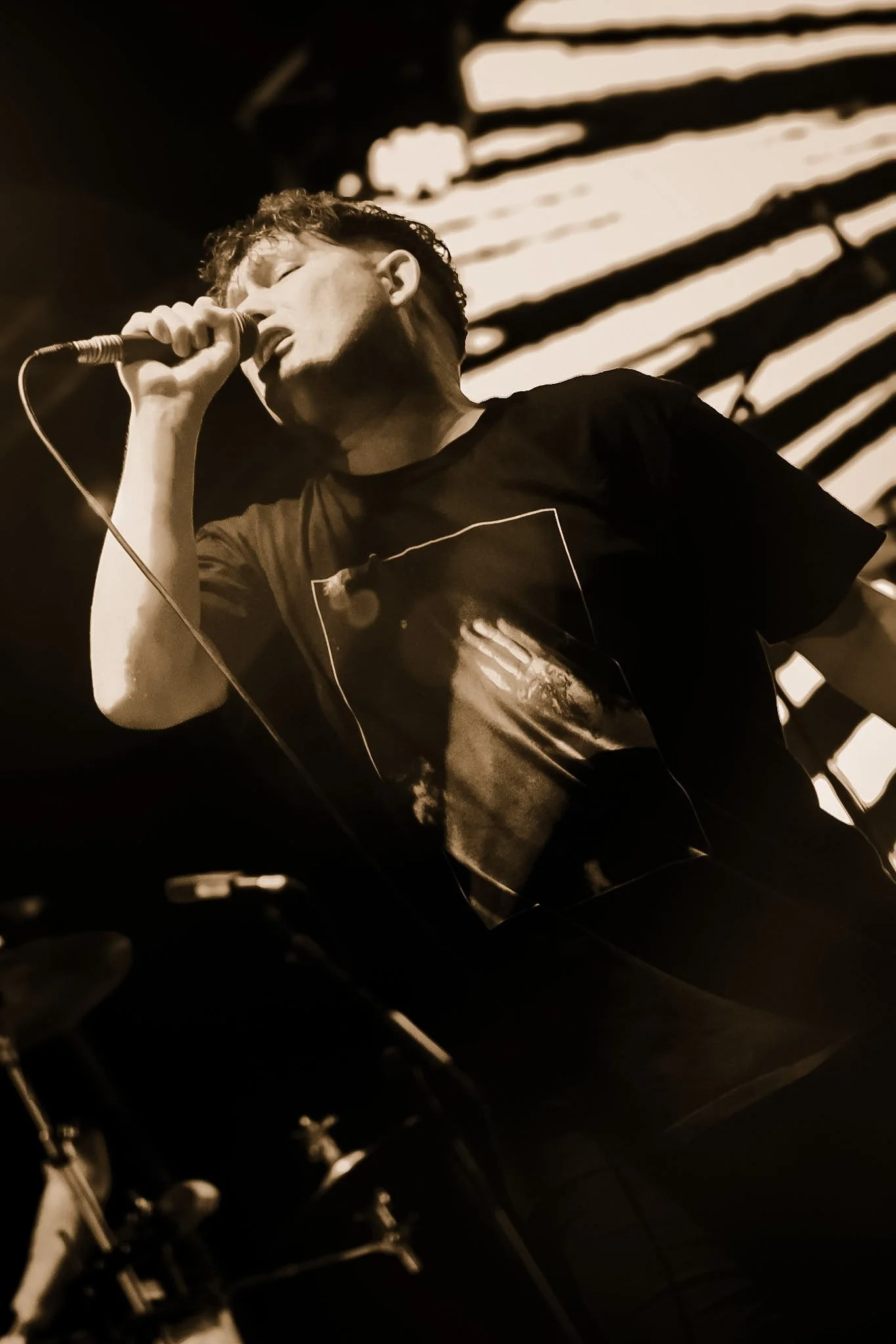 A man singing into a microphone on stage, with a background of striped light patterns.