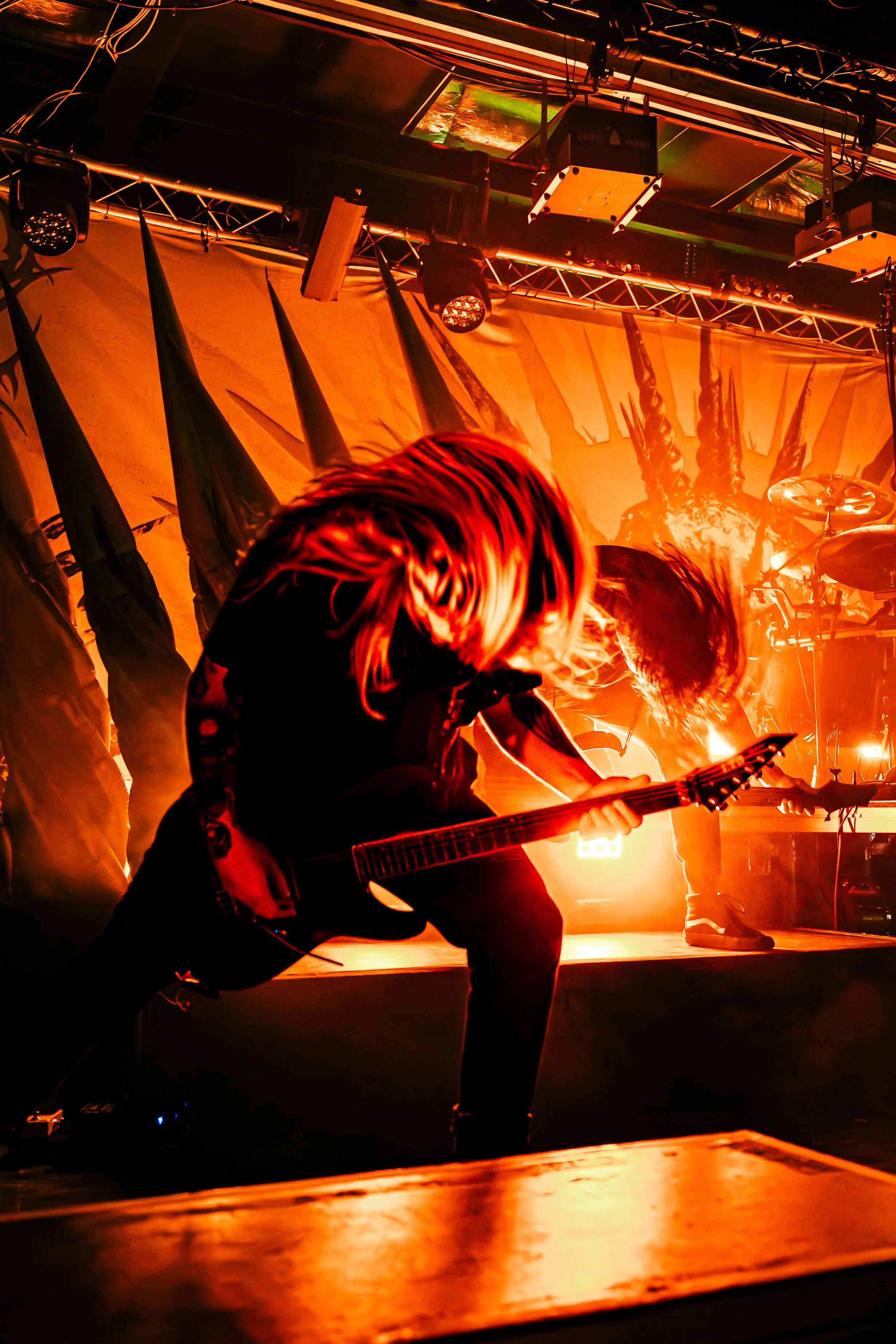 A musician is playing an electric guitar on stage, illuminated by dramatic orange and yellow stage lighting, with stage equipment and drapery visible in the background.