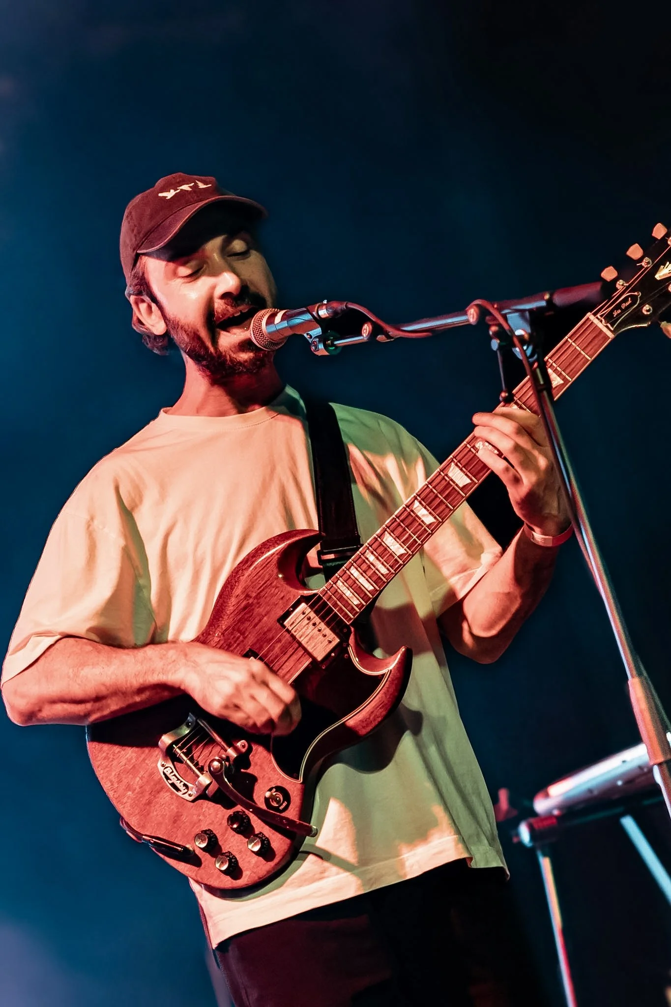 A man with a beard and mustache, wearing a baseball cap and a beige t-shirt, plays an electric guitar and sings into a microphone on stage with a dark blue background.