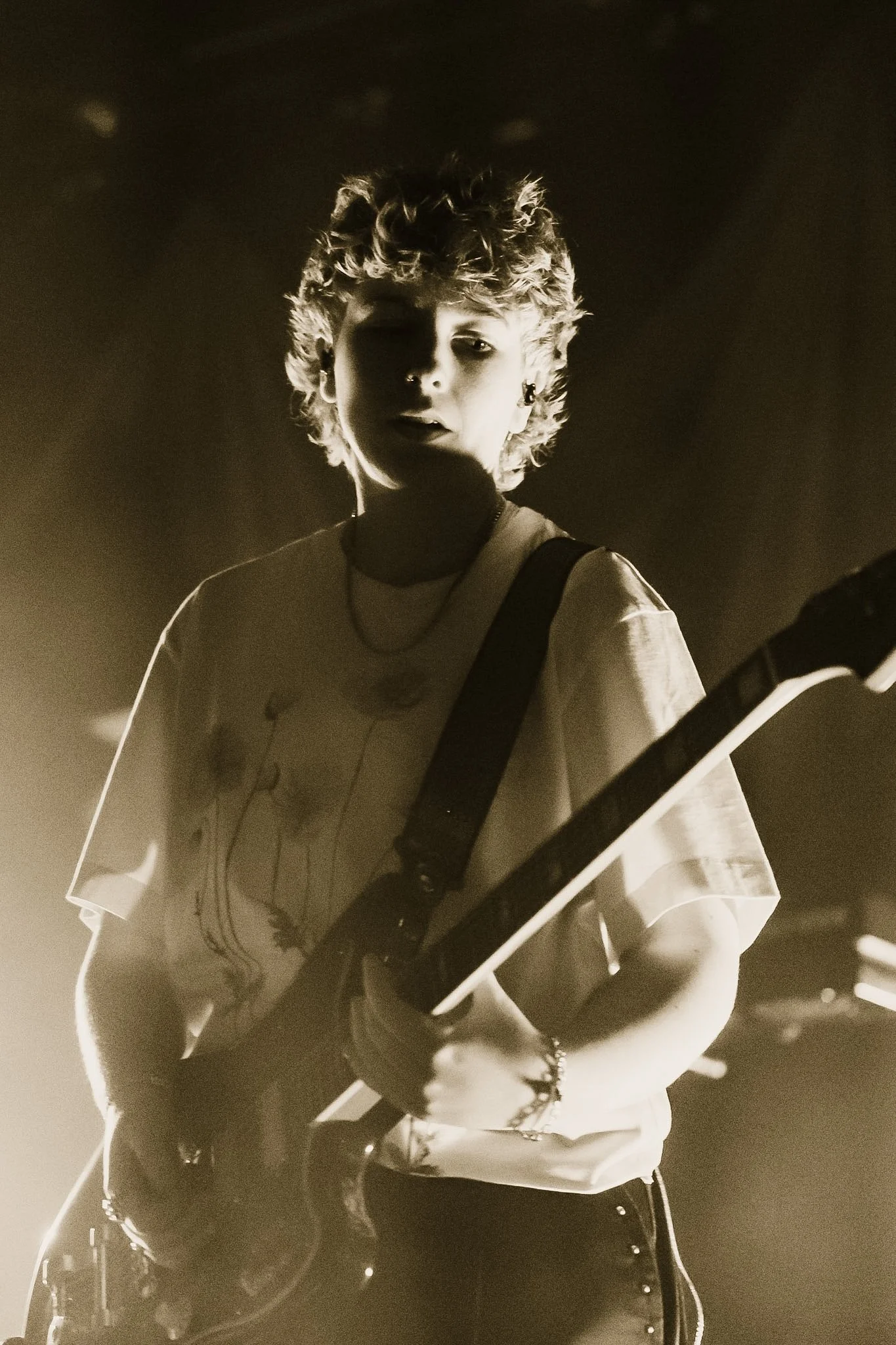 A black and white photo of a person with curly hair playing an electric guitar. They are wearing a t-shirt with a graphic print and have multiple earrings. The background is dark, and the lighting highlights the person's face and guitar.