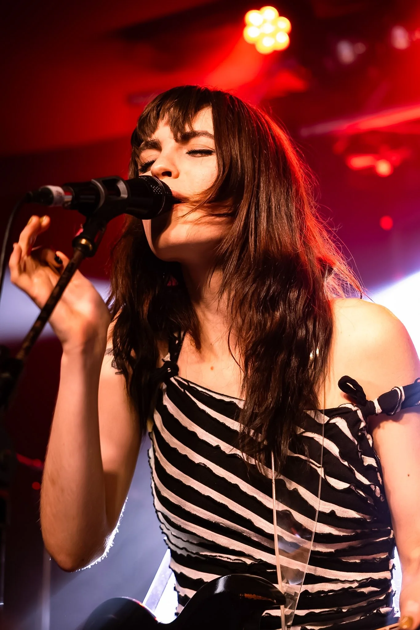 A woman with dark hair singing into a microphone on stage with red lighting.