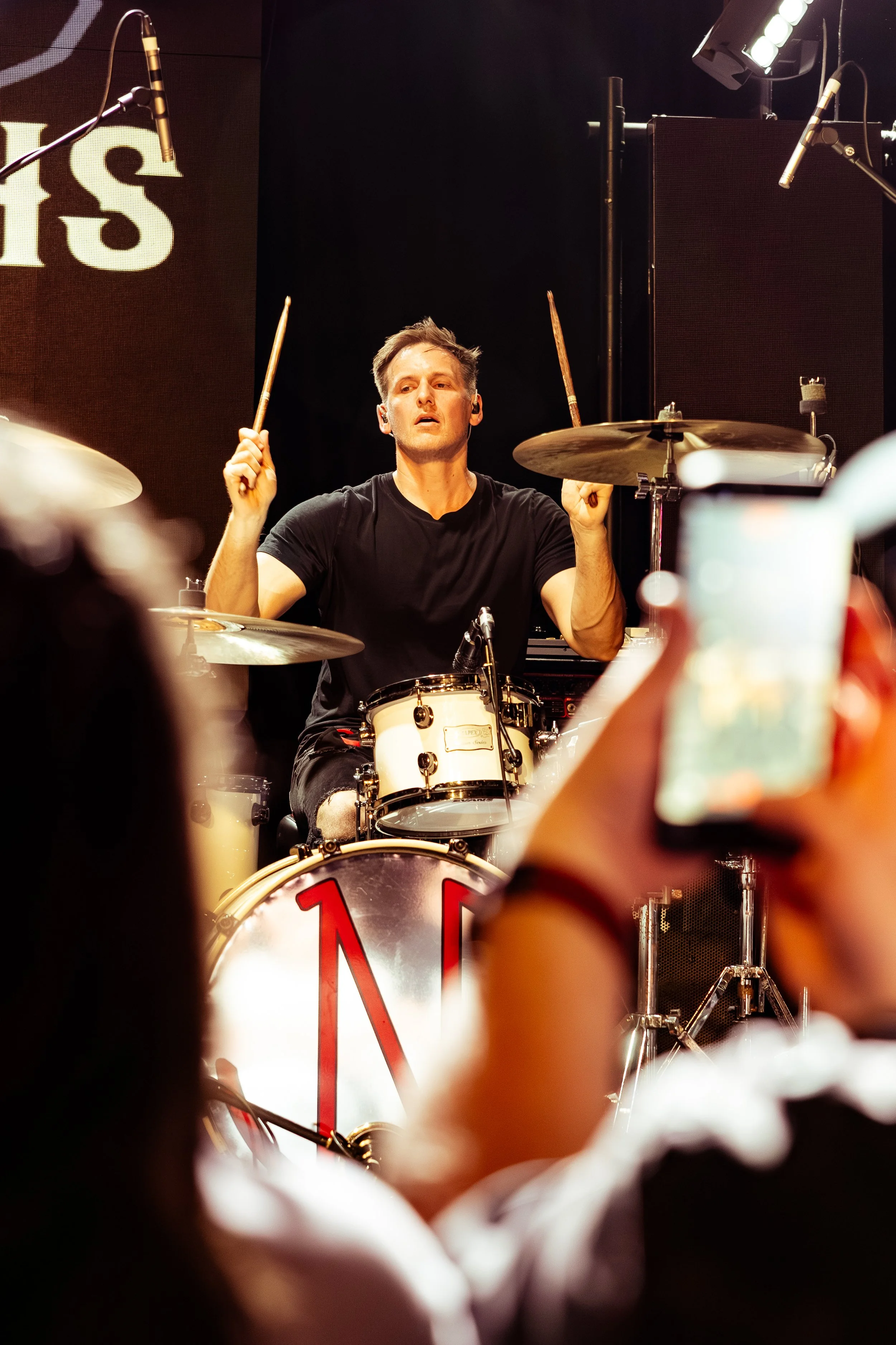 A male drummer performs on stage, surrounded by musical equipment and lights, with an audience capturing the moment on their smartphones.