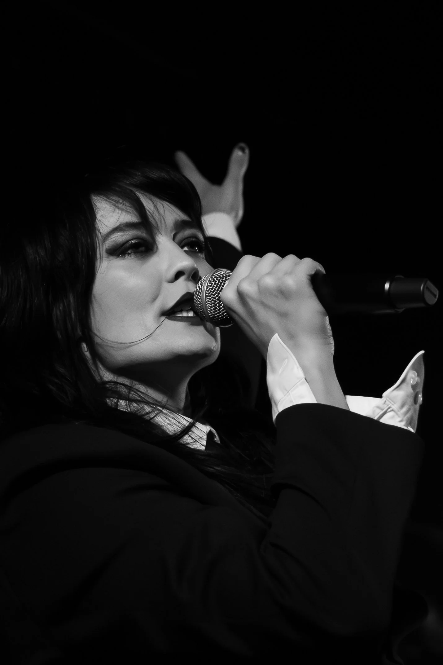 A woman with dark hair and makeup is singing into a microphone, with her eyes partially closed and her hand raised, in a black and white photo.