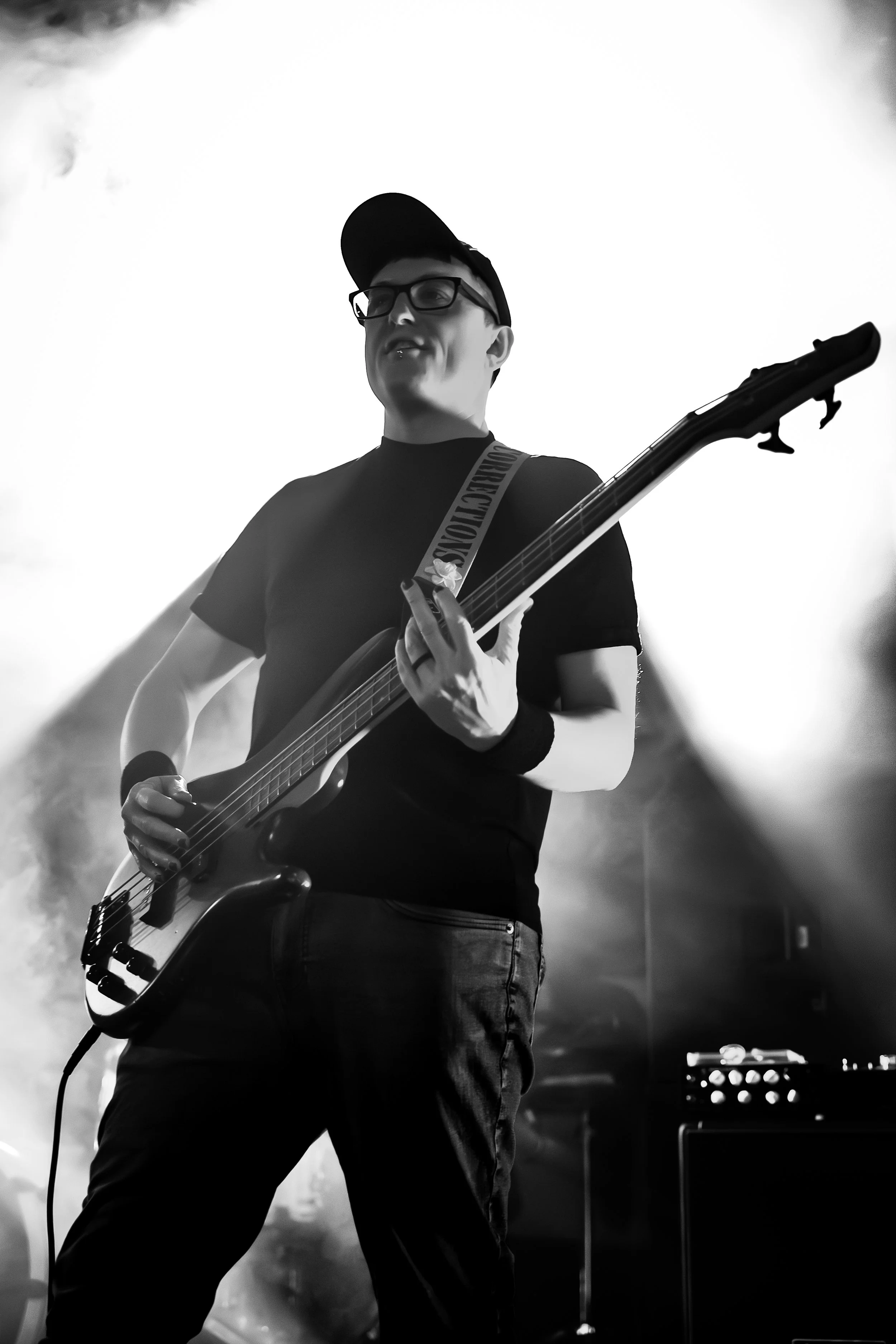A person wearing a baseball cap and glasses playing an electric guitar on stage amidst smoke and stage lights.
