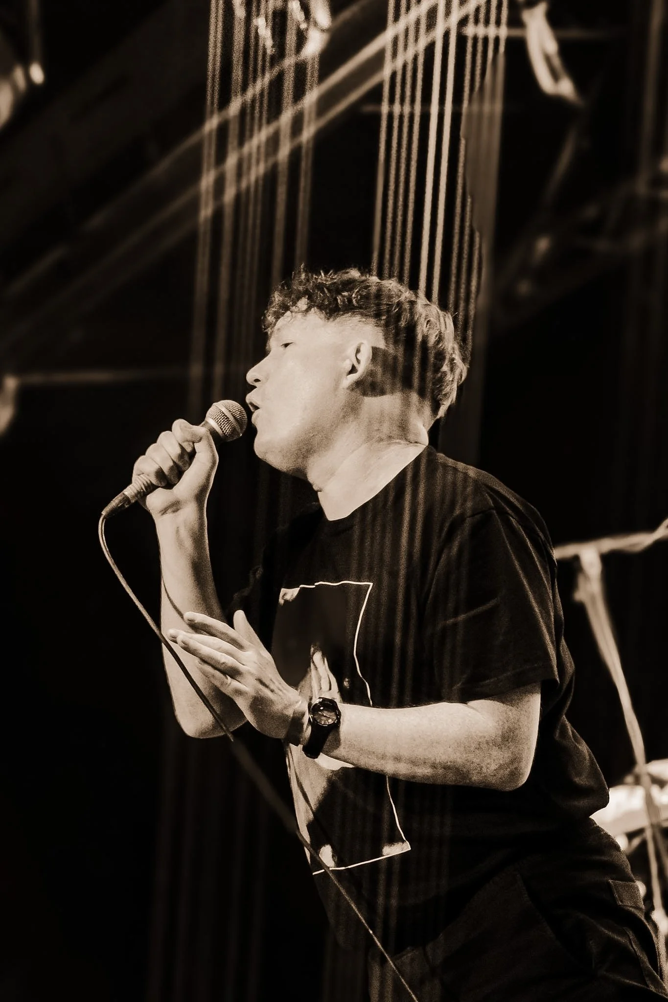 A young man is performing on stage, singing into a microphone with eyes closed and one hand raised, with a dark background and stage lighting.