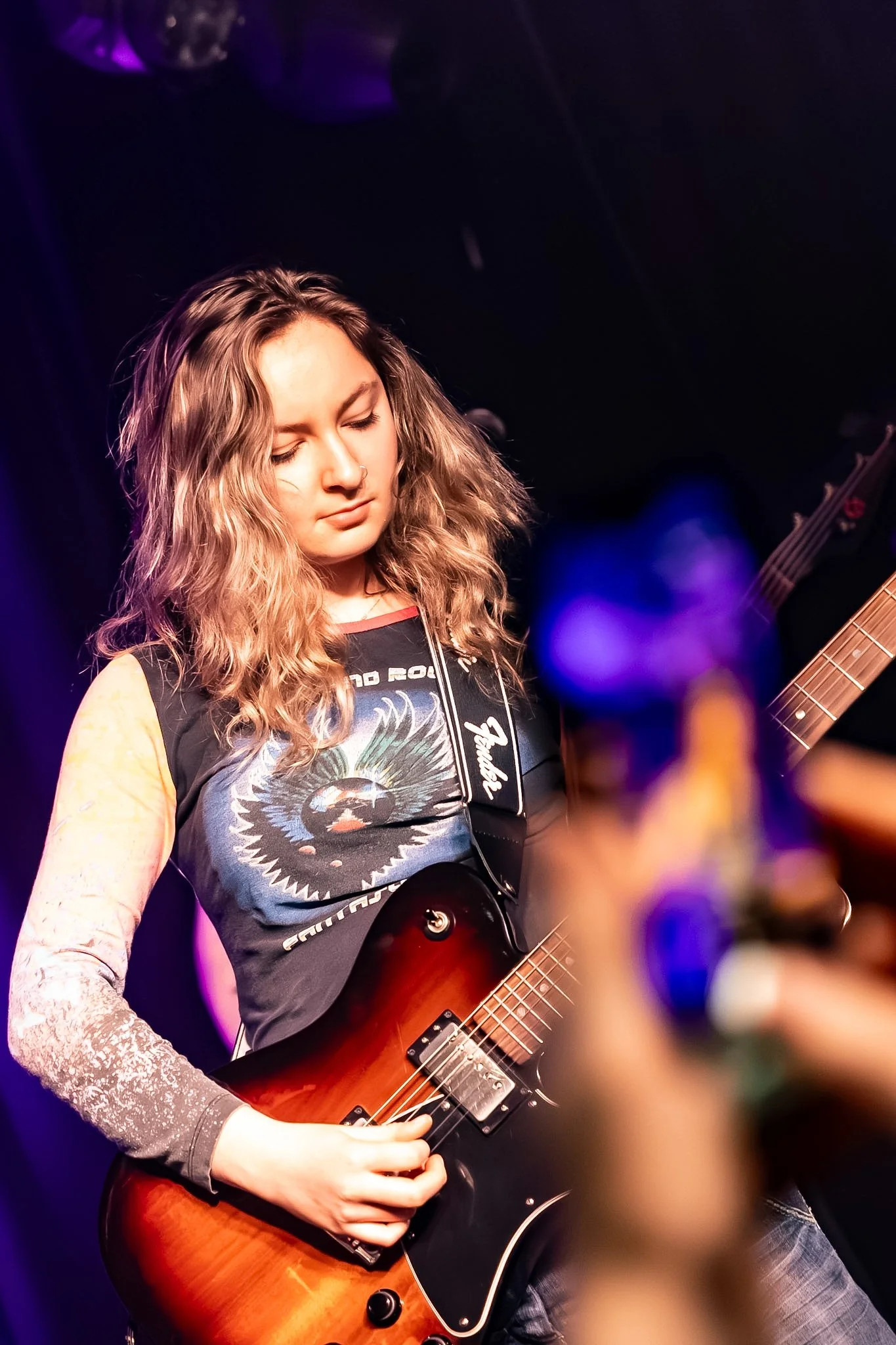 A young woman with wavy blonde hair plays an electric guitar on stage, wearing a sleeveless shirt with a graphic design, and standing under purple stage lighting.