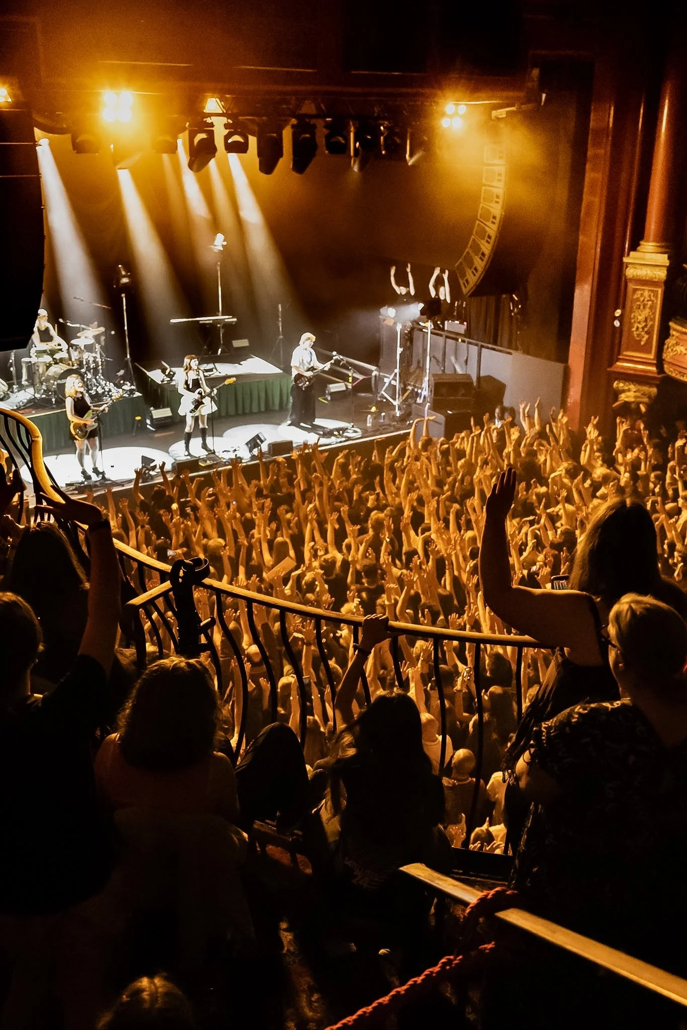 Concert stage with bright yellow stage lights, band members performing, and an audience with raised hands in a theater or concert hall.