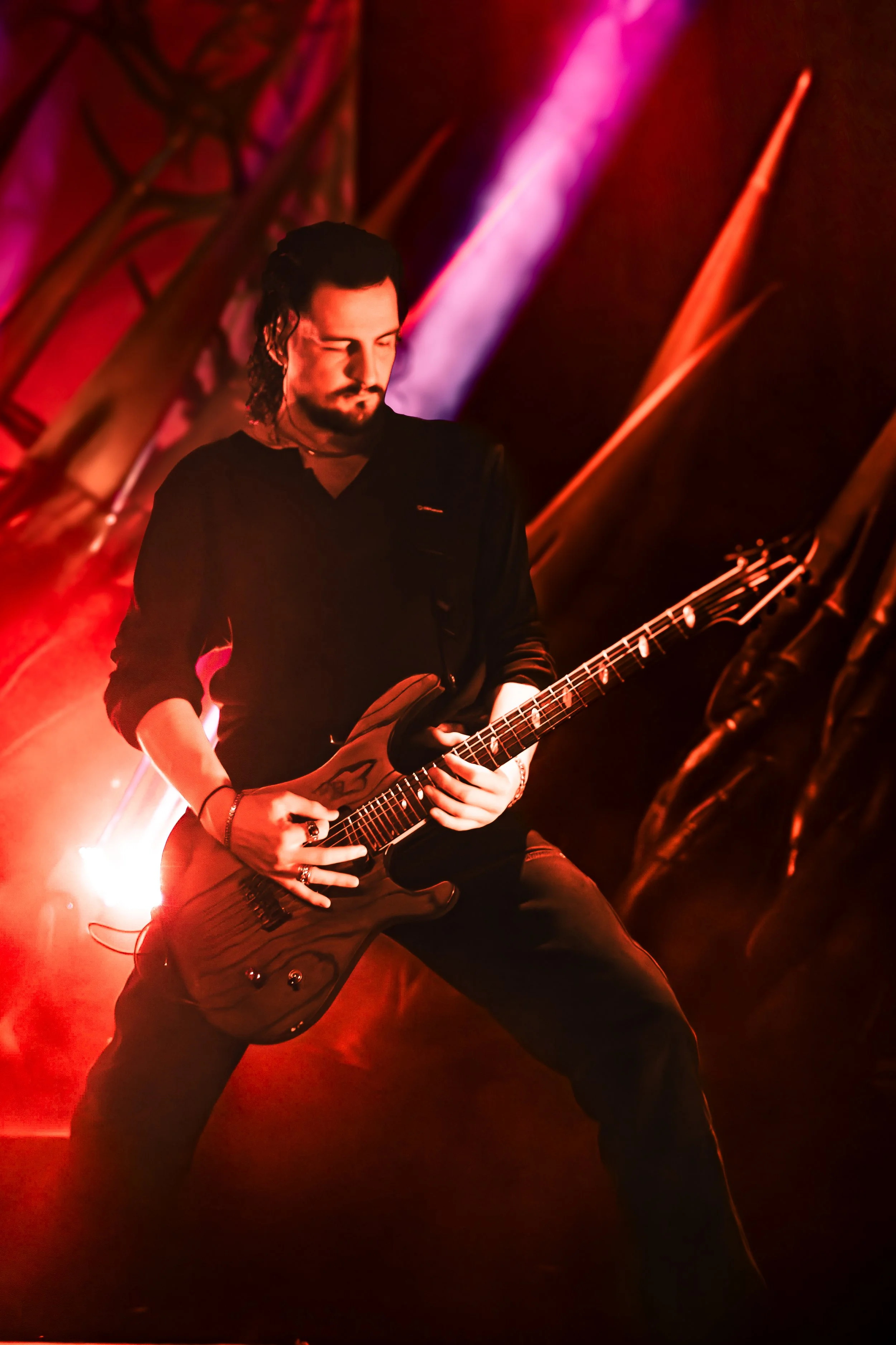 A musician with long dark hair and a beard playing an electric guitar on stage with red and purple lighting.
