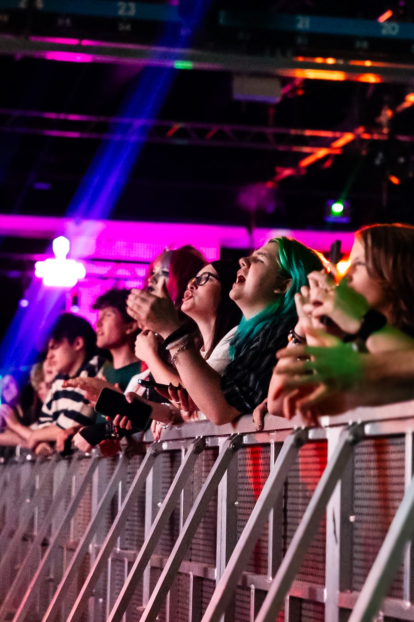 People at a concert or event, standing behind a metal barrier with colorful lights and a digital display in the background.