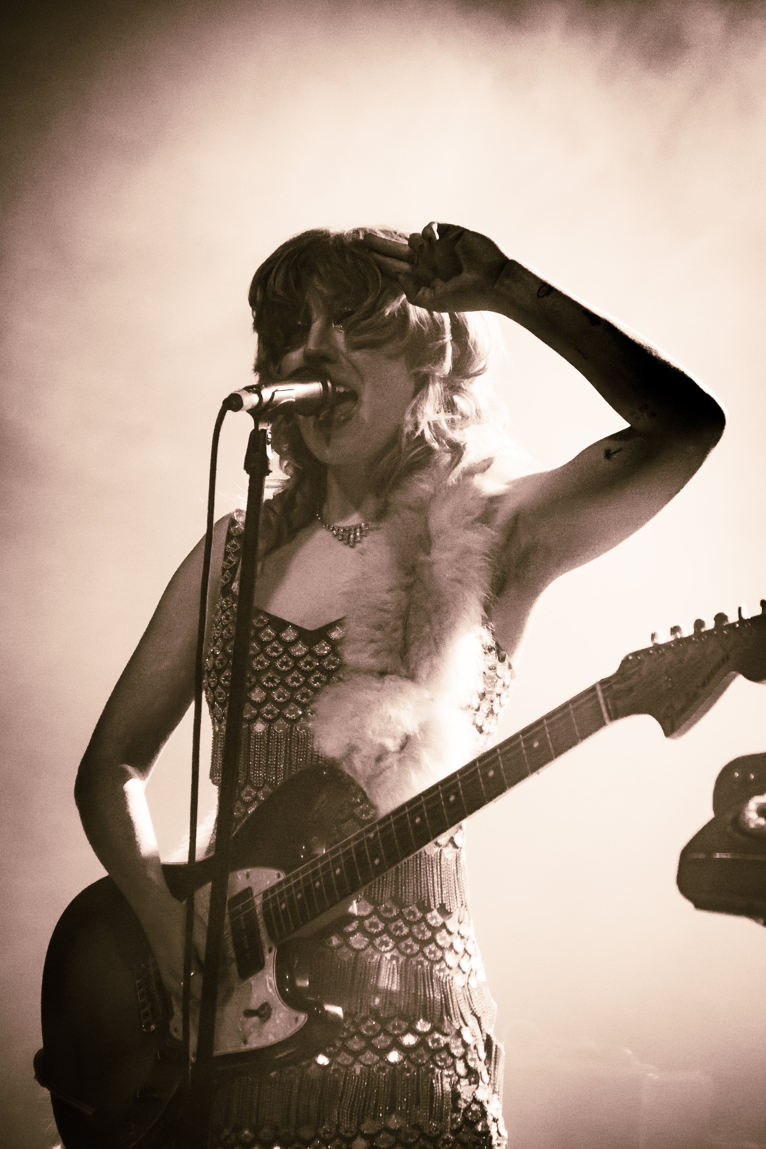 A woman singing into a microphone while playing an acoustic guitar, wearing a shiny dress and a furry stole, with a spotlight behind her creating a silhouette effect.