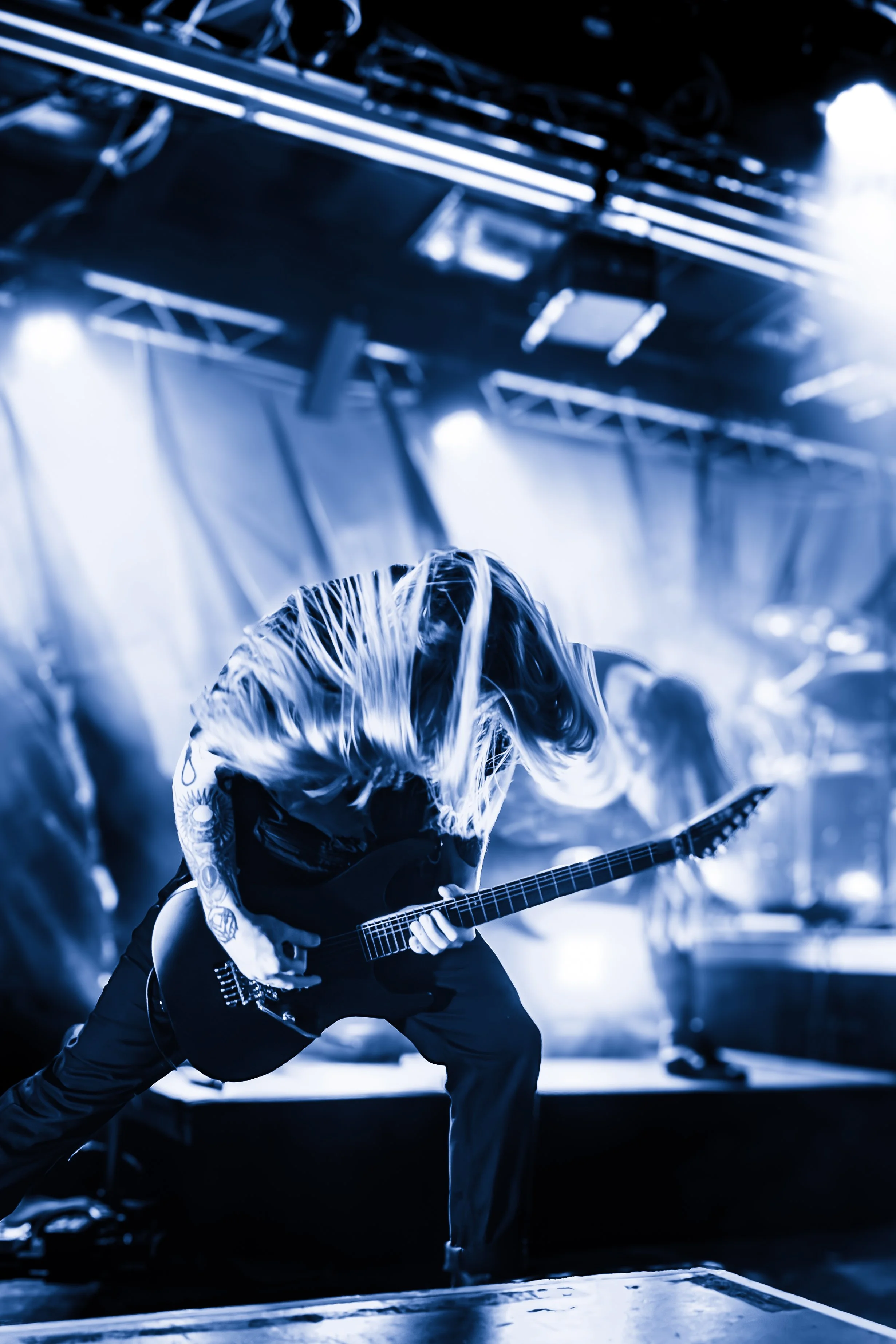 A musician with long hair playing an electric guitar on stage, illuminated by bright stage lights in a dark venue.