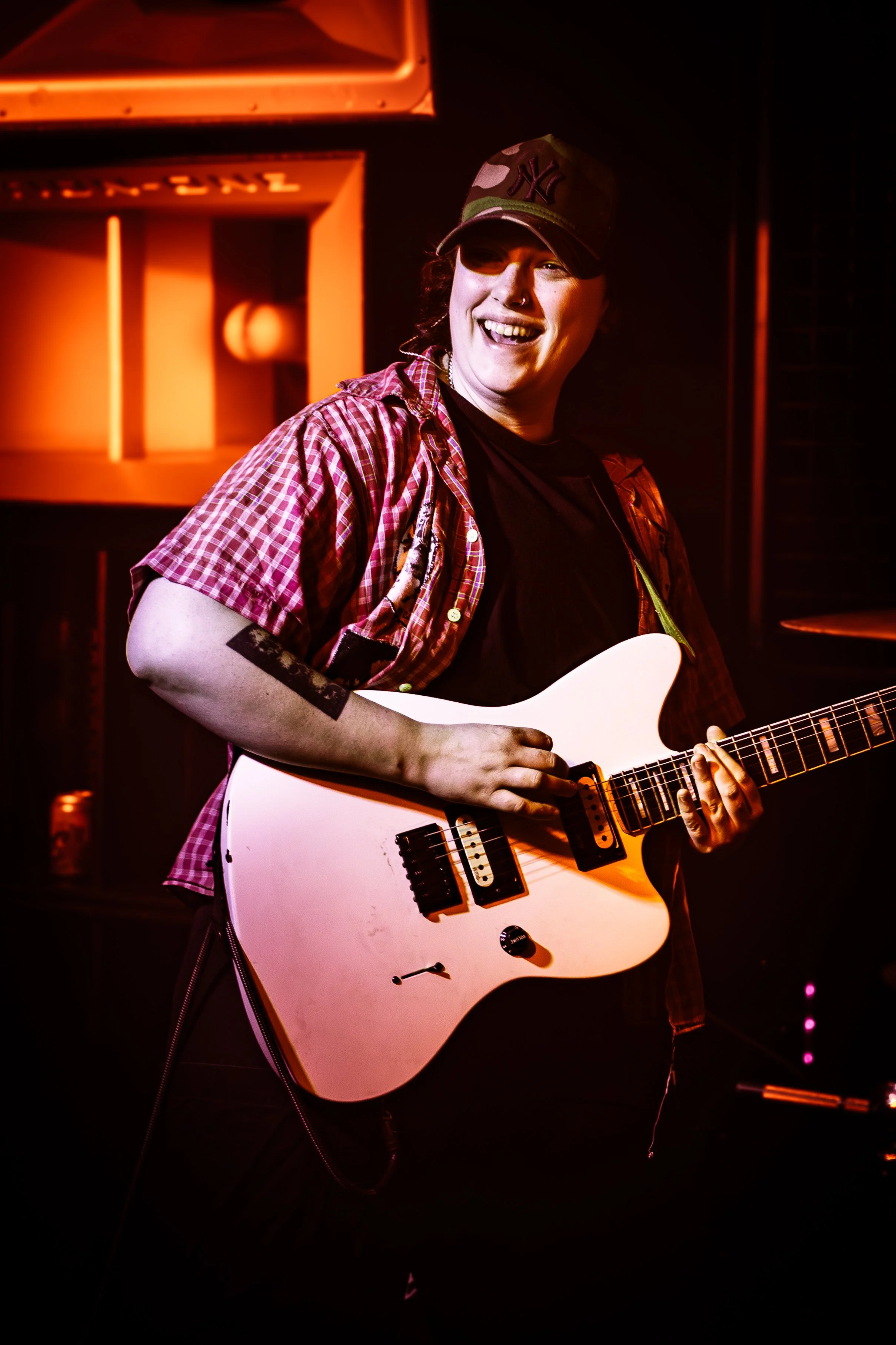 A young musician smiling and playing a white electric guitar on stage, wearing a camouflage cap and a red checkered shirt, illuminated by warm stage lighting.