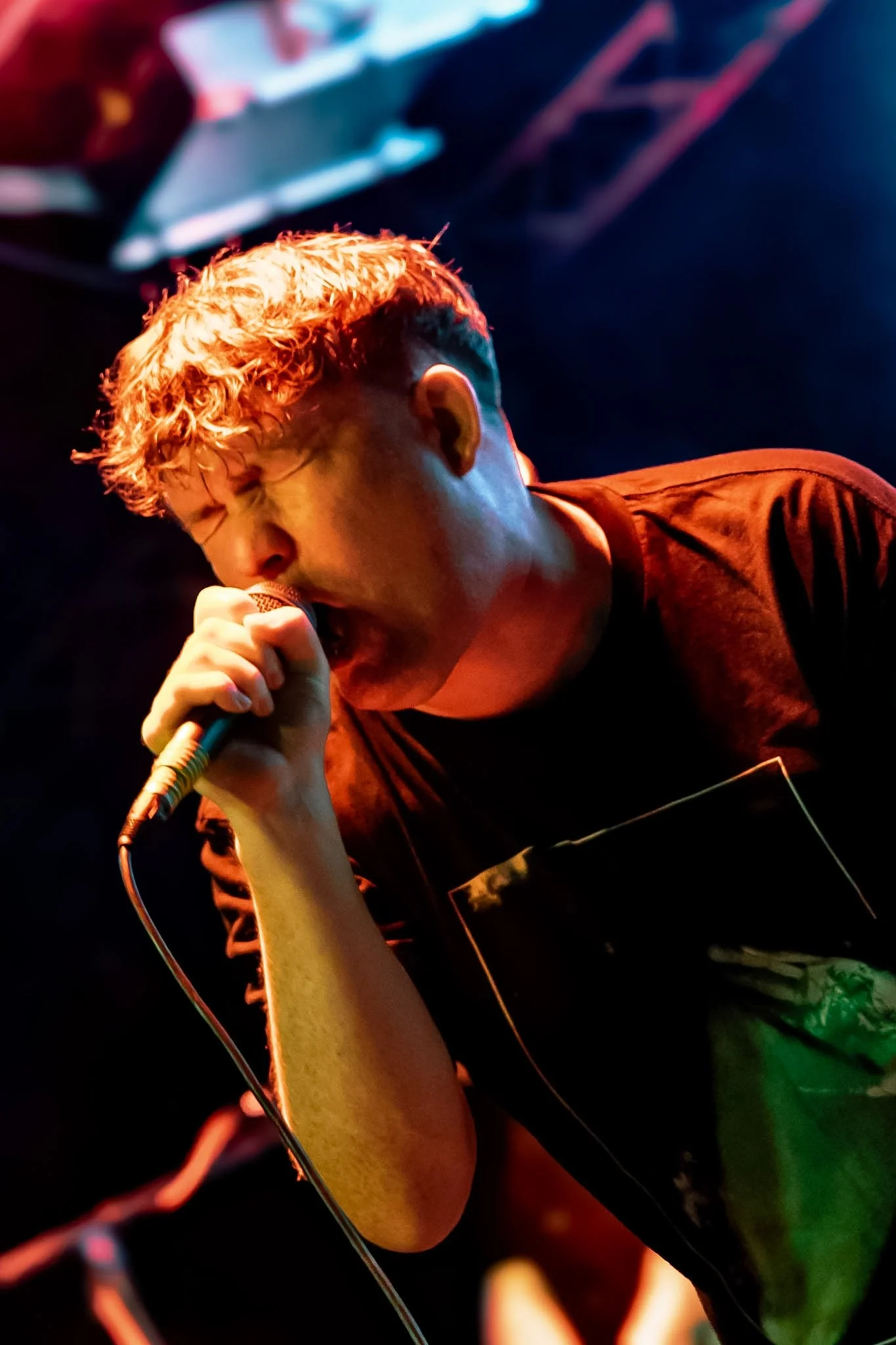 Close-up of a person singing into a microphone during a performance, with curly blonde hair illuminated by stage lighting, wearing a black t-shirt, and a blurred background with vibrant colors.