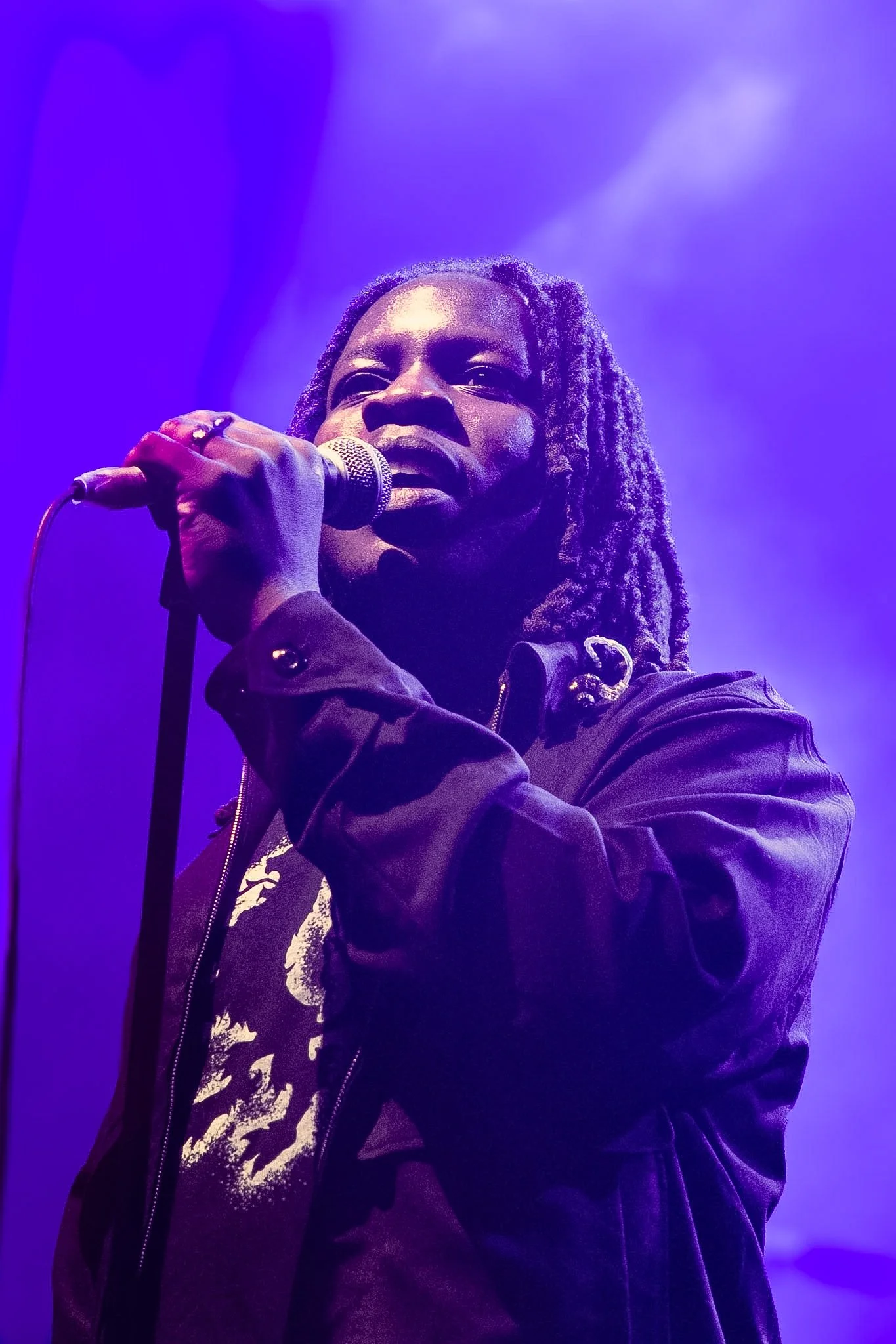 A person with dreadlocks holding a microphone, wearing a dark jacket, under purple stage lighting.