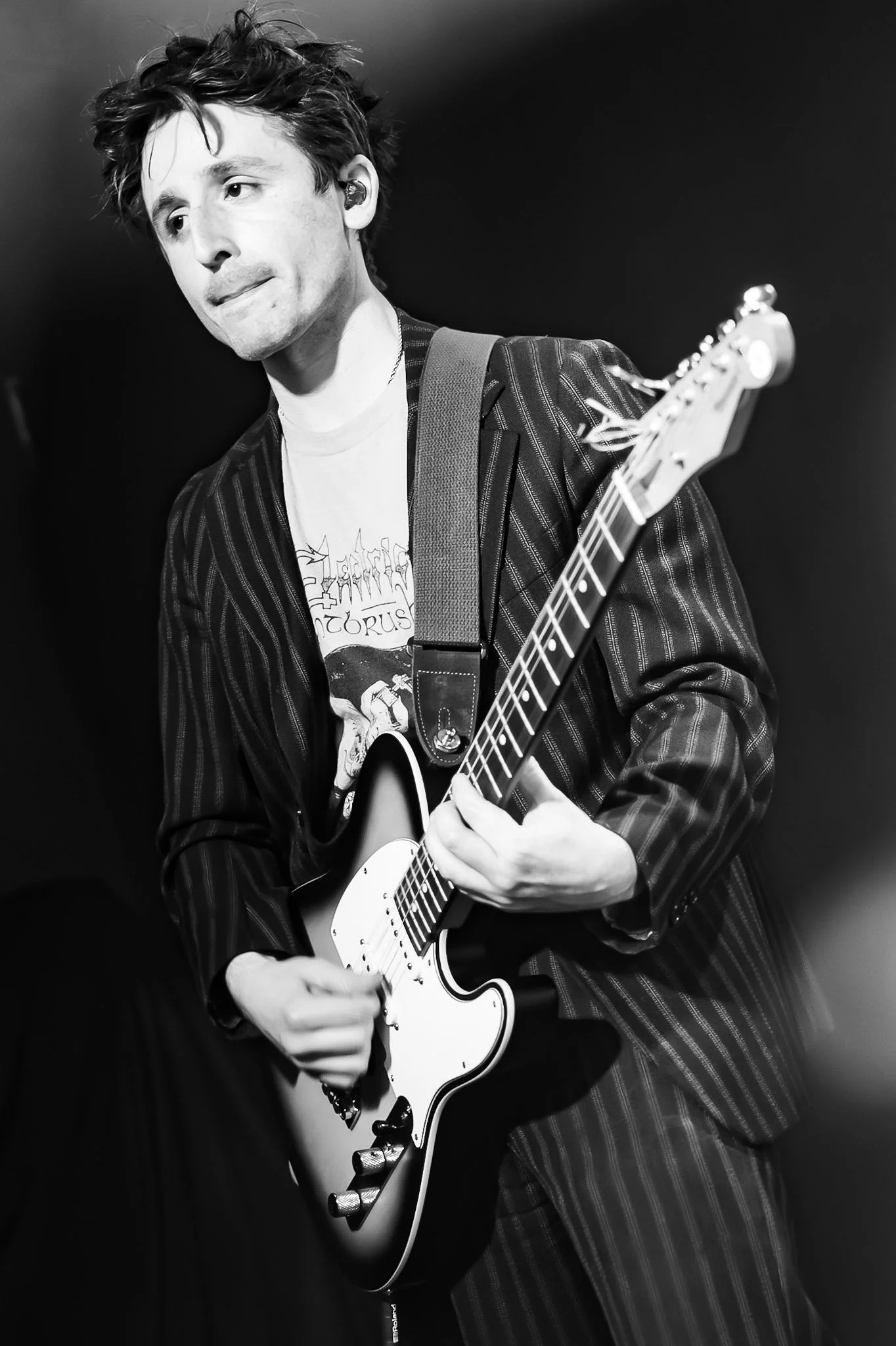 A young man with messy hair playing an electric guitar on stage, wearing a striped blazer and a graphic T-shirt, in black and white.
