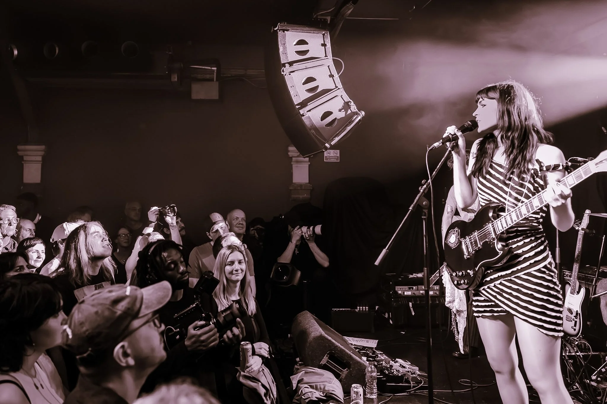 A female musician with long wavy hair performing on stage, playing an electric guitar and singing into a microphone, with an audience watching.