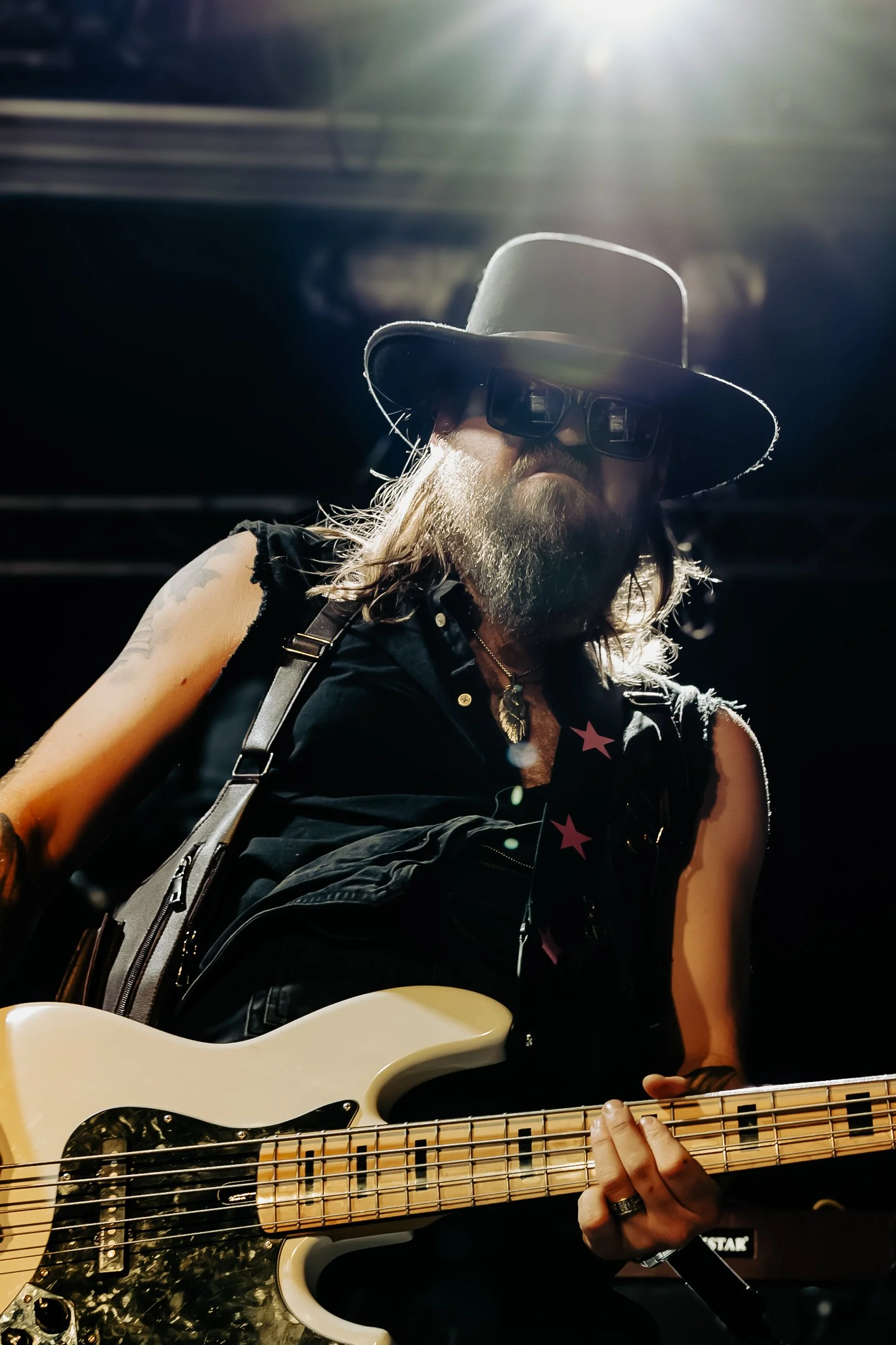 Person with long hair, a beard, wearing sunglasses, a wide-brimmed hat, a sleeveless vest with star patches, and a cross necklace, playing an electric guitar.