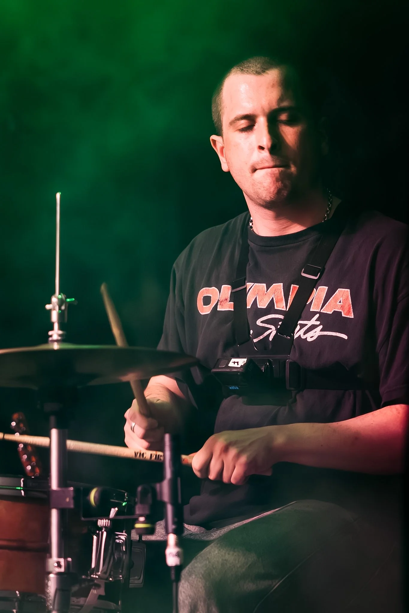 A young man with short hair playing the drums, wearing a black T-shirt with orange and white text, and a beaded necklace. He has his eyes closed and is focused on playing the drums, with musical equipment visible around him, against a dark, moody bac