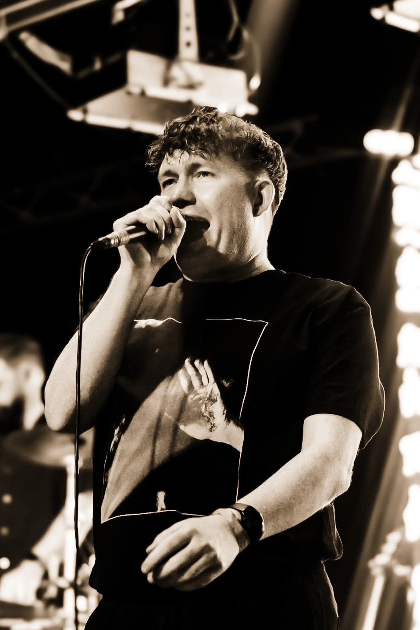 A young man with curly hair singing into a microphone in a live music venue, with stage lighting in the background.