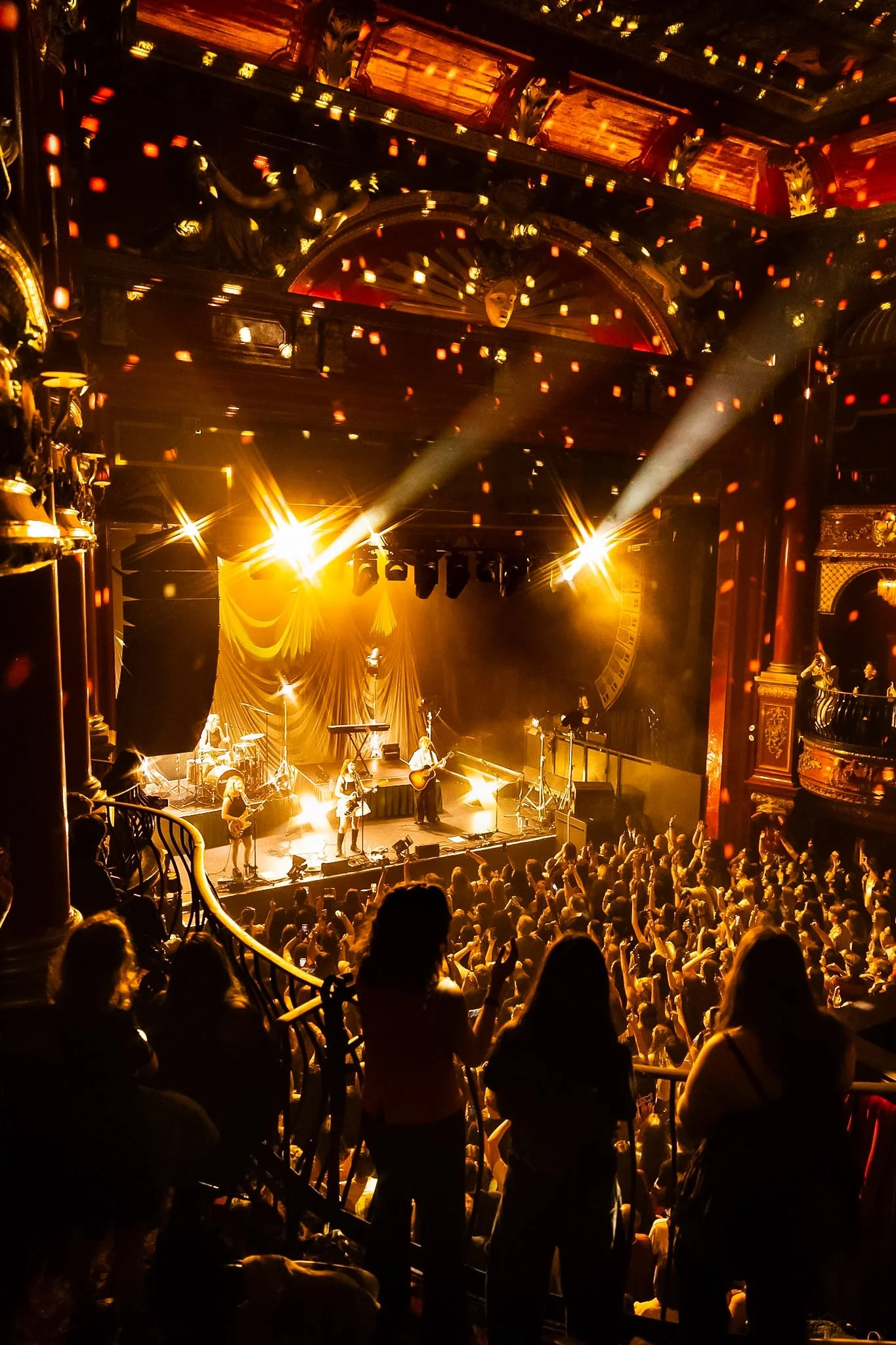 A live music concert inside a theater with a band performing on stage, bright yellow stage lighting, and an audience watching the performance.