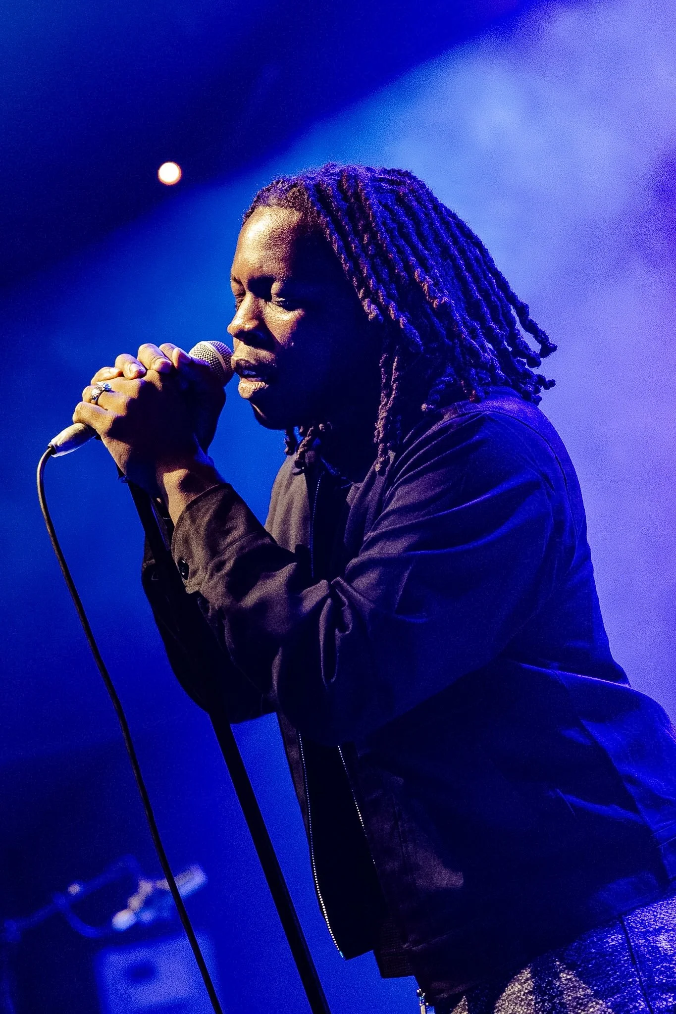 A man with long hair singing into a microphone on stage, with blue and purple stage lighting.