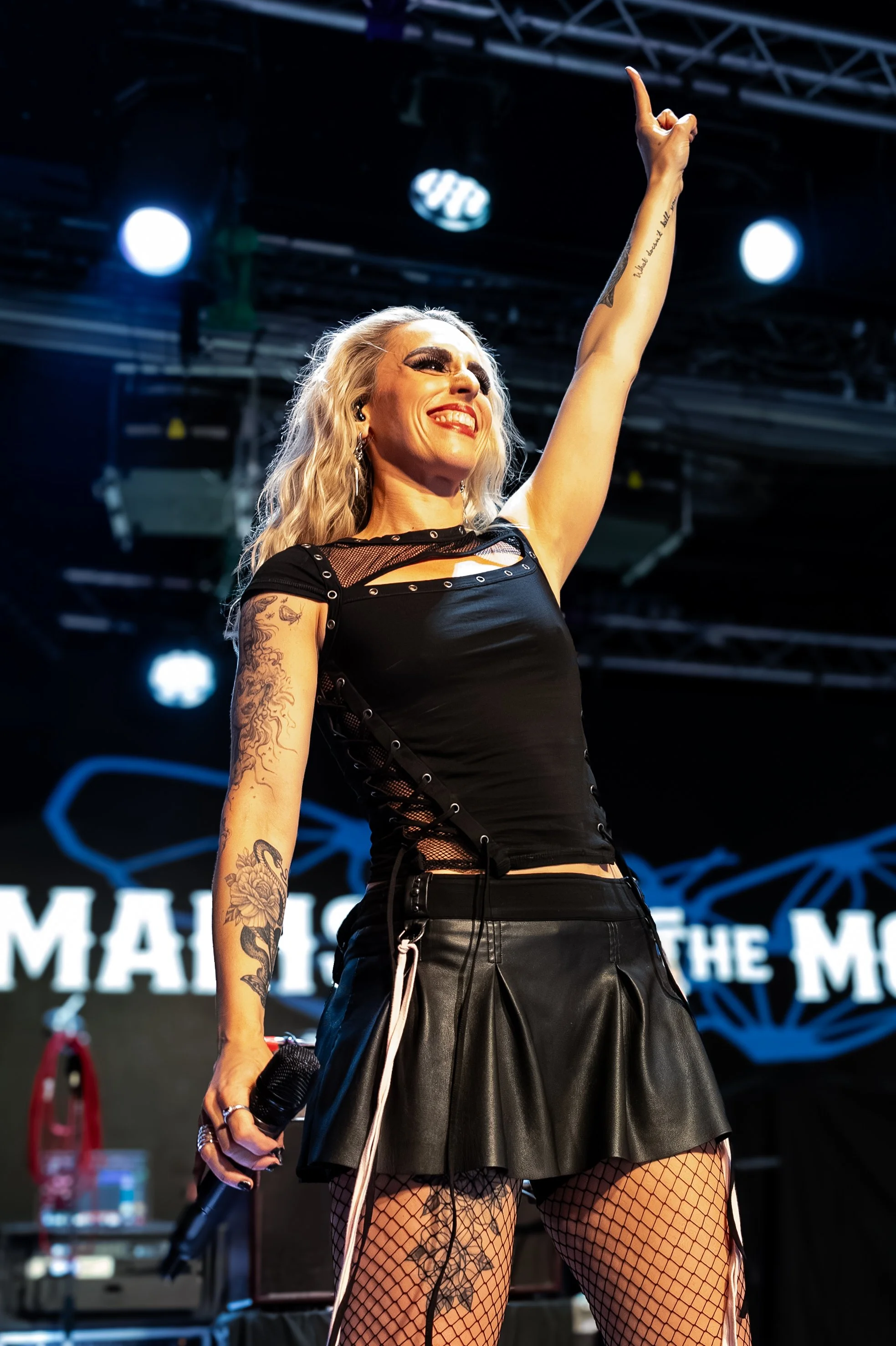 A woman with long blonde hair, tattoos, and fishnet stockings on stage, smiling and raising her right arm with her index finger pointing upward, holding a microphone in her left hand, under stage lights.