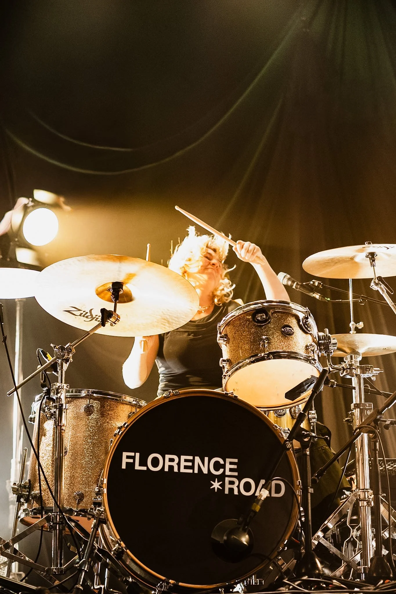 A drummer with curly blonde hair is playing a drum kit on stage. The drum kit has a bass drum with the words "FLORENCE + THE ROLLING STONES". The drummer is holding drumsticks and is surrounded by cymbals, drums, and microphone stands.